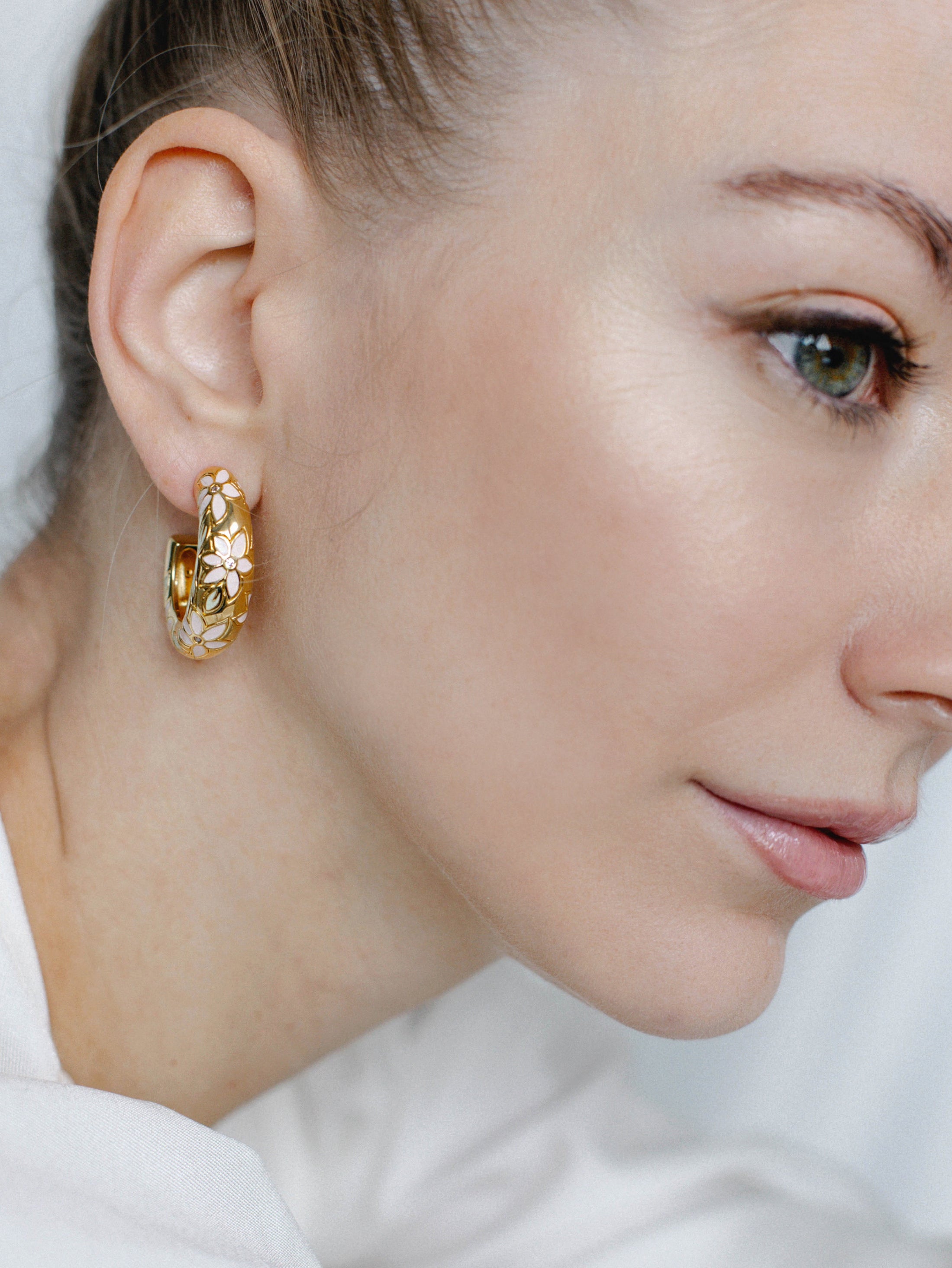 A close-up image of a woman's ear wearing a gold-toned floral hoop earring with hand-painted pastel flowers and crystal accents.