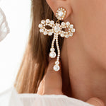 A pair of dangling crystal earrings with gold plating, designed in the shape of bows, adorning a person's ear.