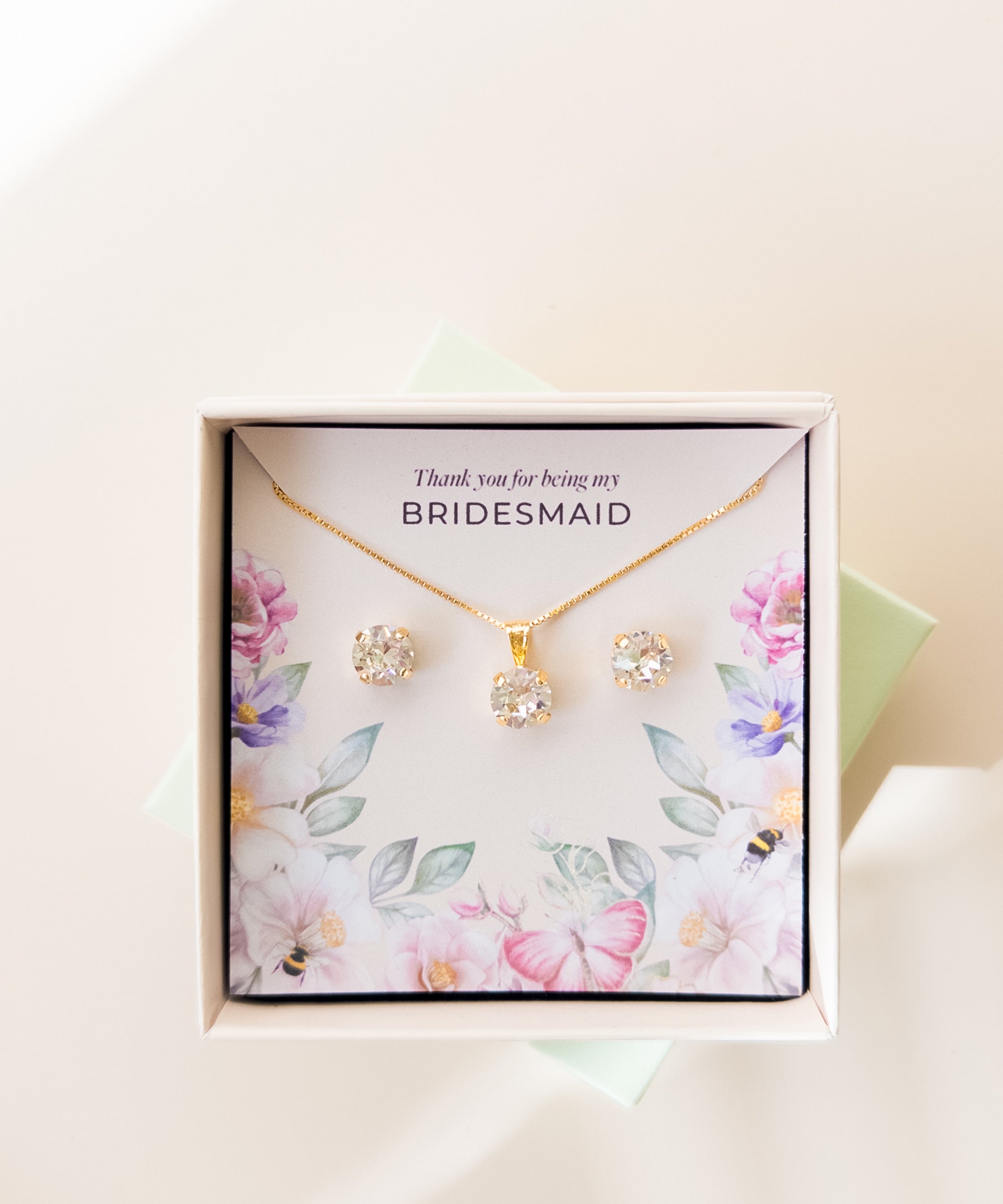 A jewelry set including a necklace and stud earrings, displayed in a box with a floral pattern and the text 'Thank you for being my Bridesmaid'. The jewelry is plated in gold, and has gold and white crystals.