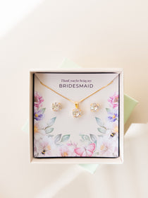 A jewelry set including a necklace and stud earrings, displayed in a box with a floral pattern and the text 'Thank you for being my Bridesmaid'. The jewelry is plated in gold, and has gold and white crystals.