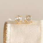 A pair of gold-plated or rhodium-plated earrings with a halo of crystals below each stud, displayed on a beige cushion.