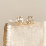 A pair of gold-plated or rhodium-plated earrings with a halo of crystals below each stud, displayed on a beige cushion.