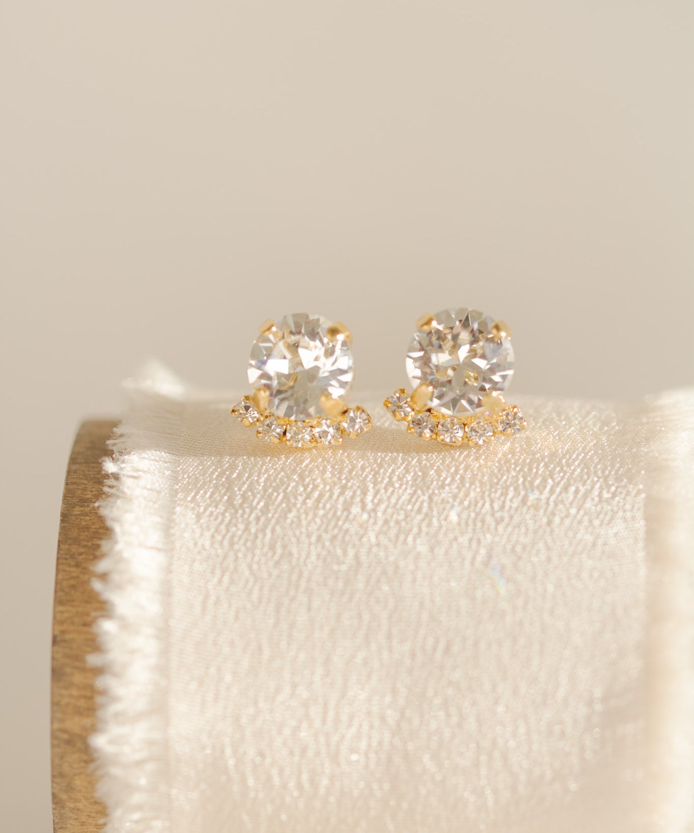 A pair of gold-plated or rhodium-plated earrings with a halo of crystals below each stud, displayed on a beige cushion.