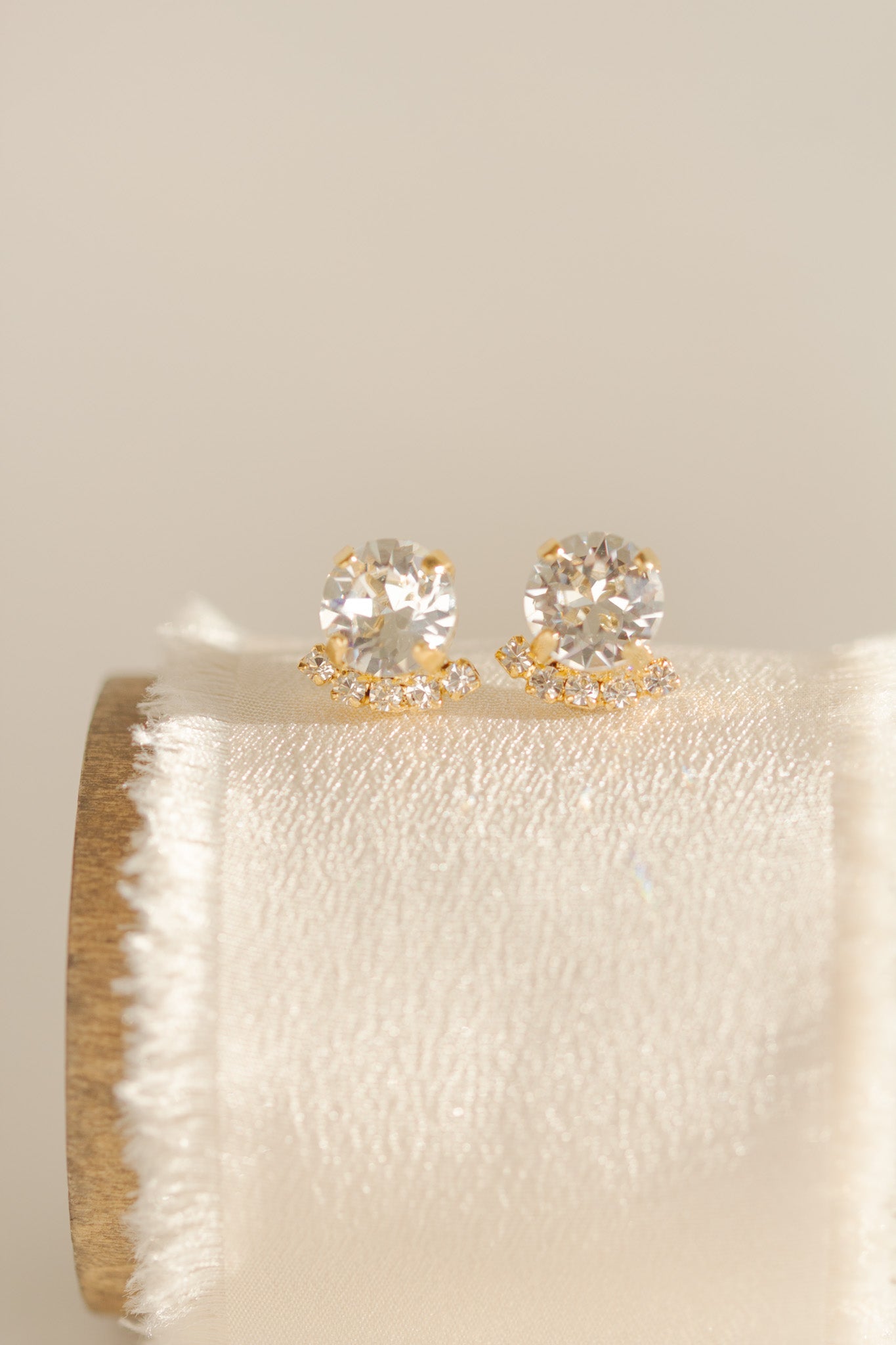 A pair of gold-plated or rhodium-plated earrings with a halo of crystals below each stud, displayed on a beige cushion.