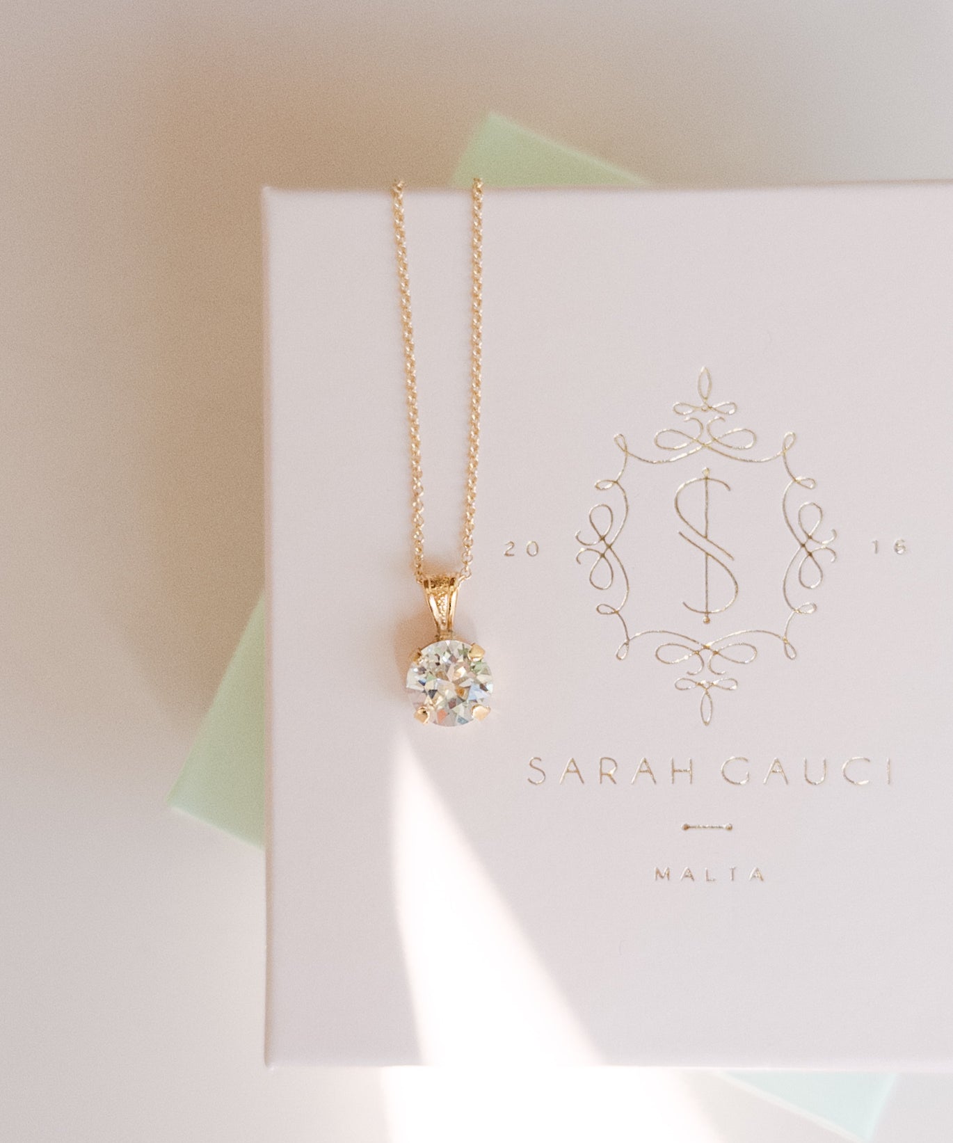 A gold-plated necklace with a crystal pendant, presented on a white box with 'SARAH GAUCI MALTA' branding.