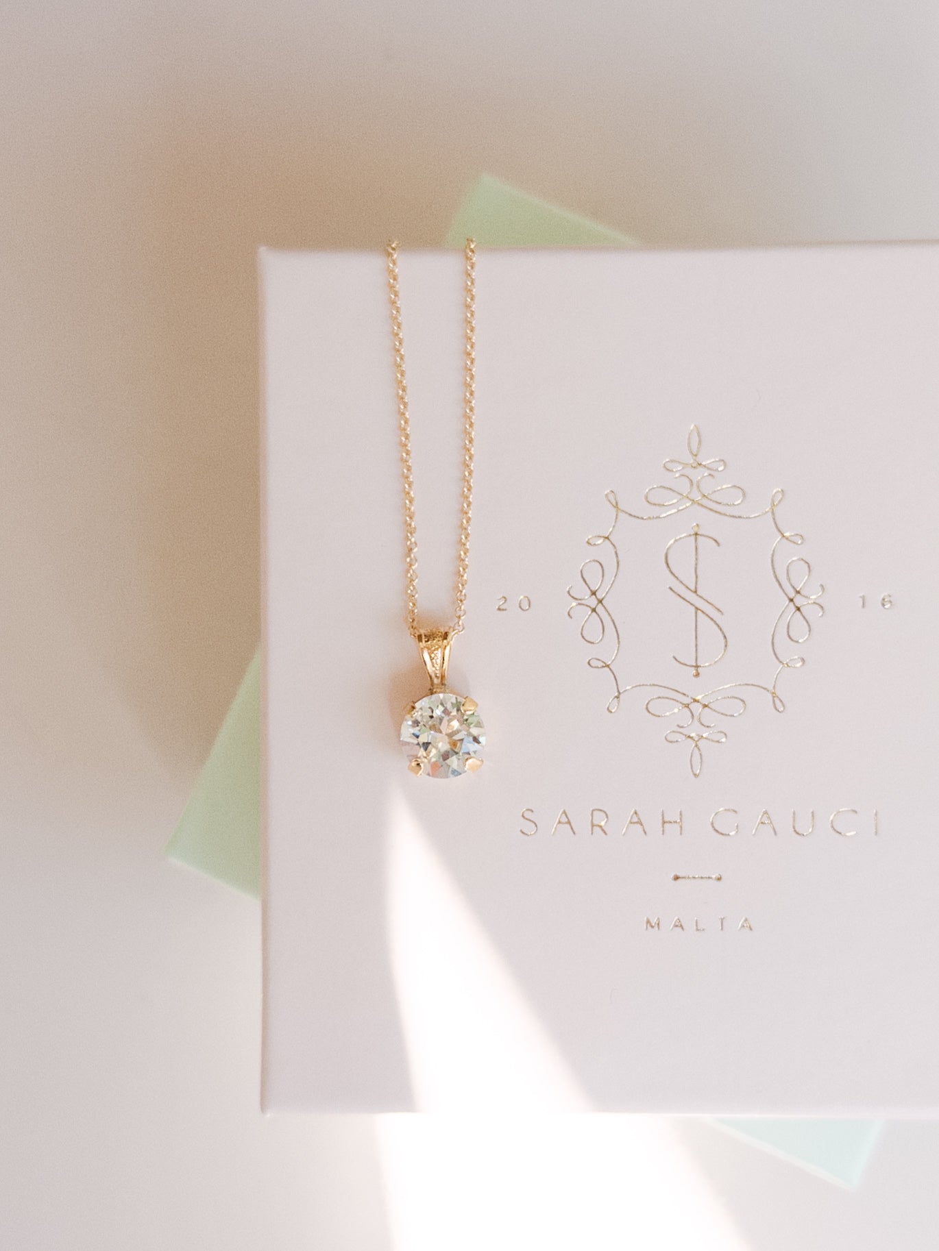 A gold-plated necklace with a crystal pendant, presented on a white box with 'SARAH GAUCI MALTA' branding.
