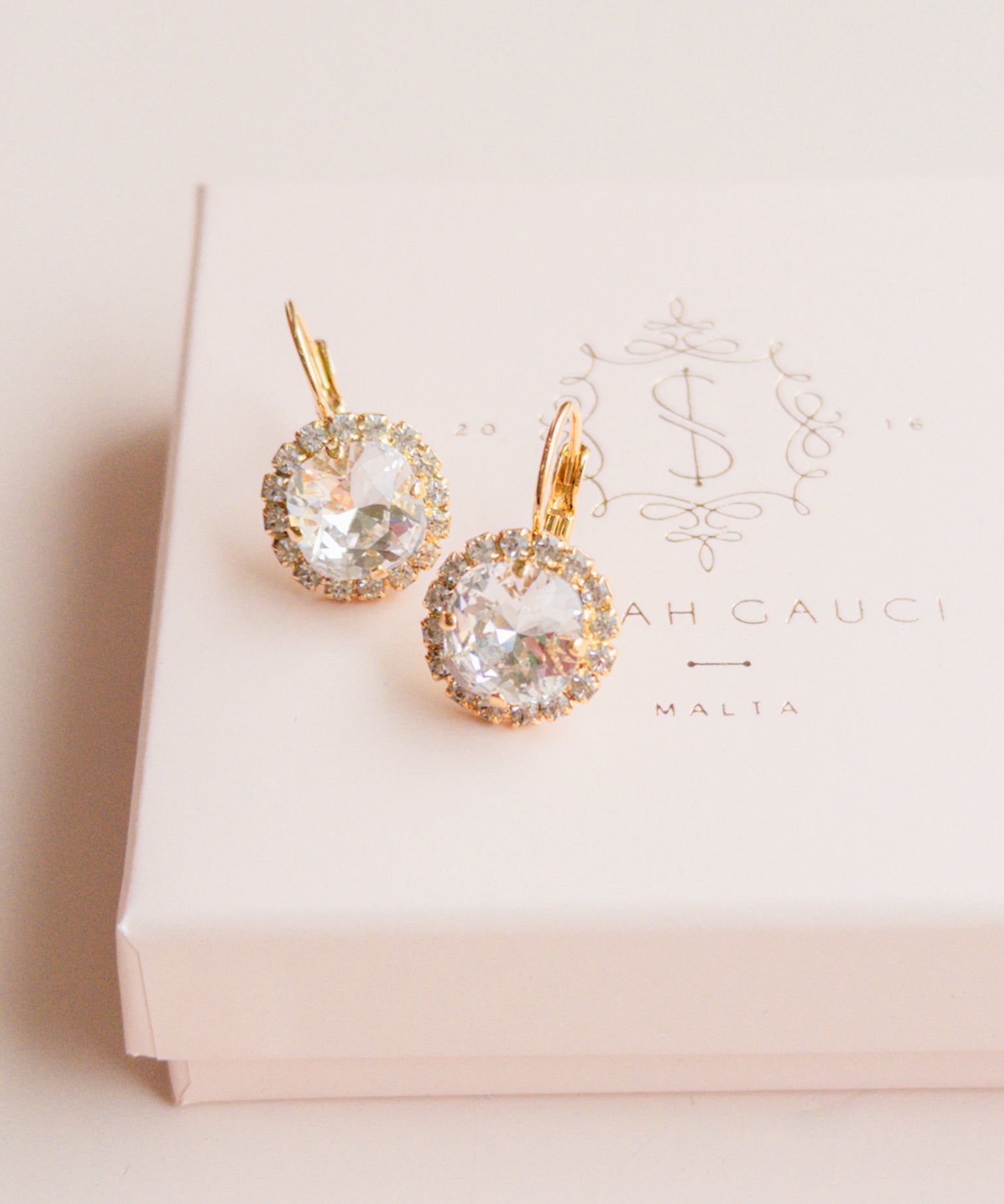 Gold earrings with clear gemstones on a pink box by Sarah Gauci Jewelry Malta 