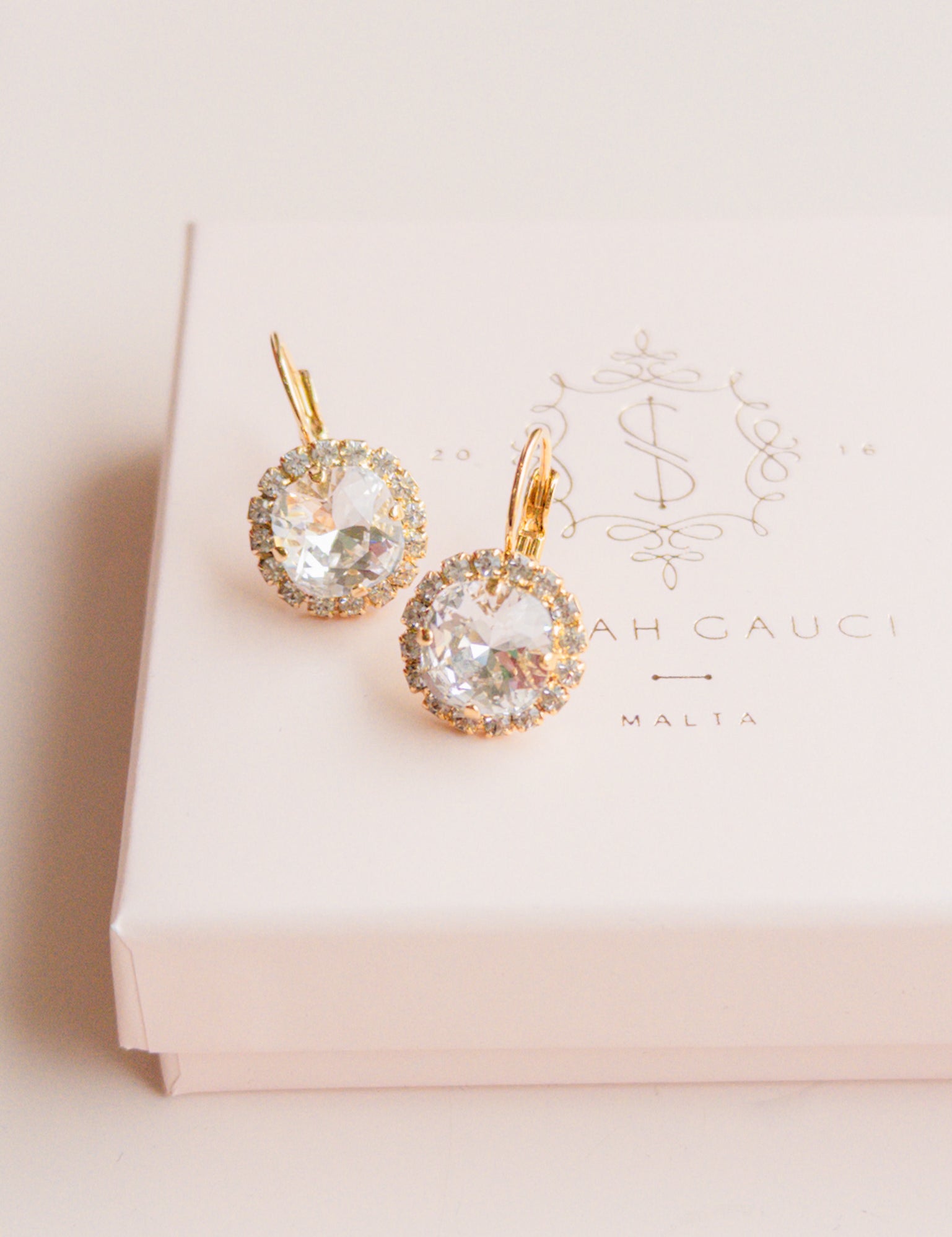 Gold earrings with clear gemstones on a pink box by Sarah Gauci Jewelry Malta 