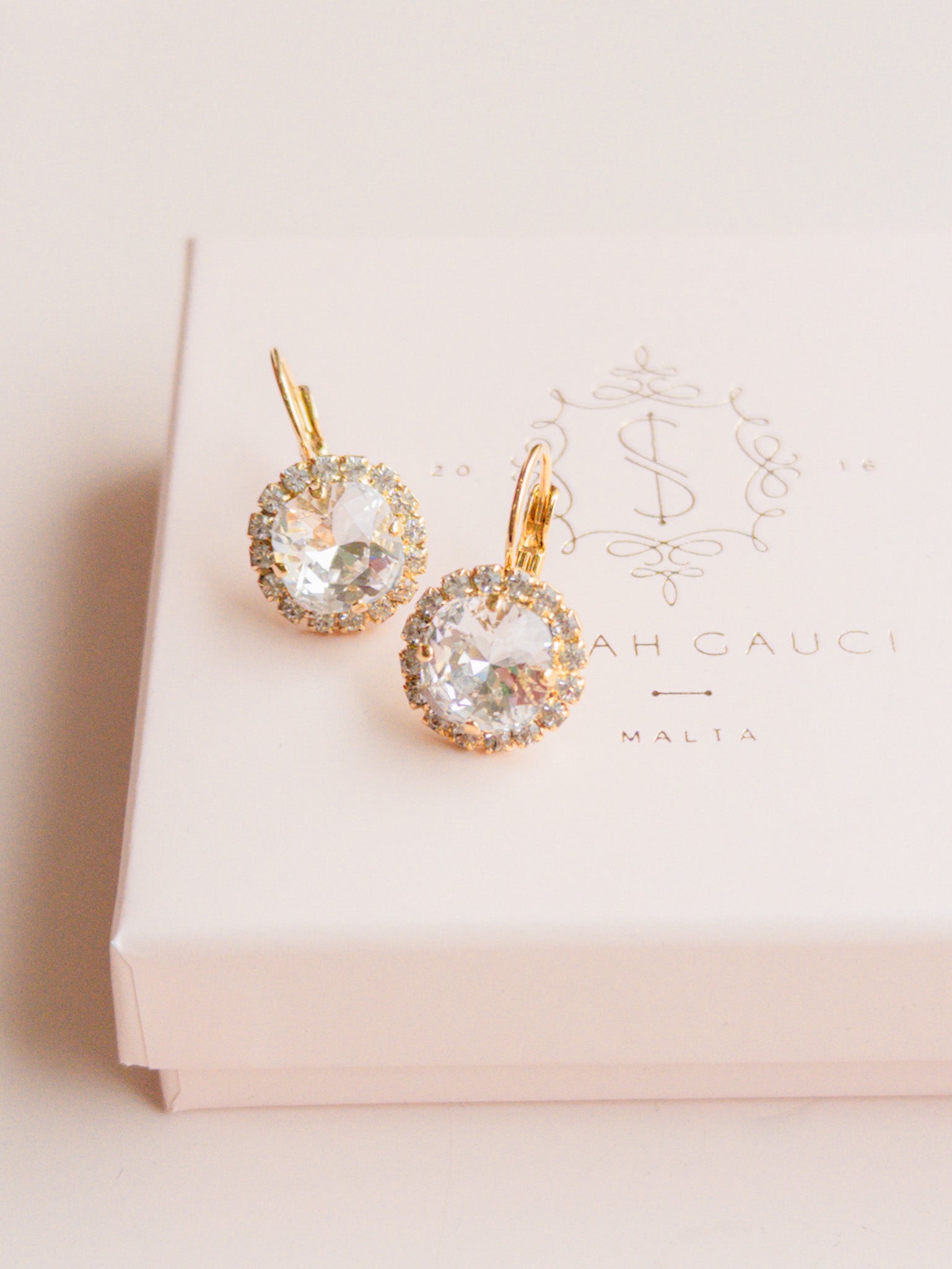 Gold earrings with clear gemstones on a pink box by Sarah Gauci Jewelry Malta 
