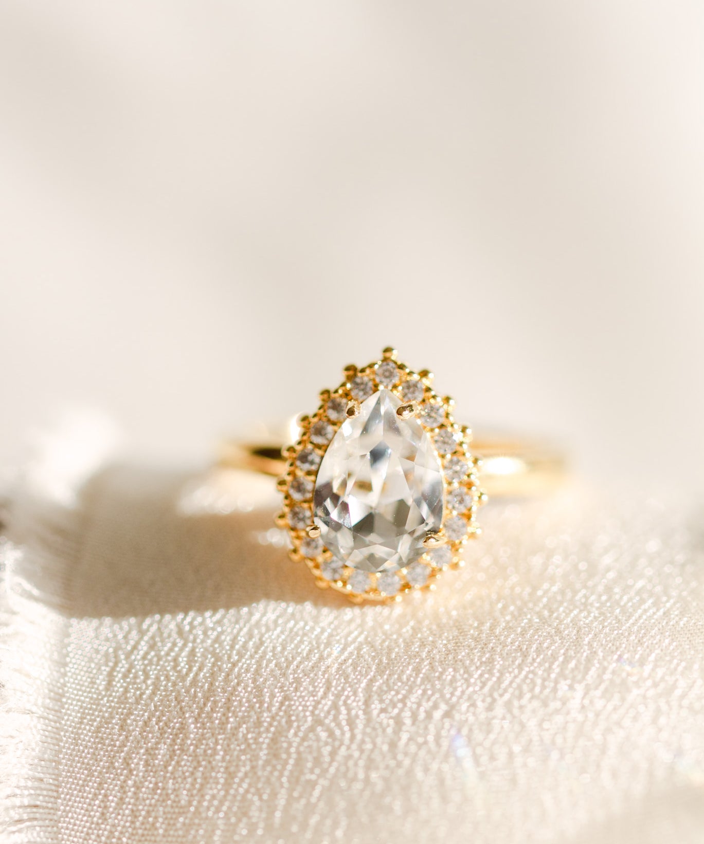 A gold-colored band ring with a central clear stone surrounded by smaller crystals, presented on a white fabric surface.