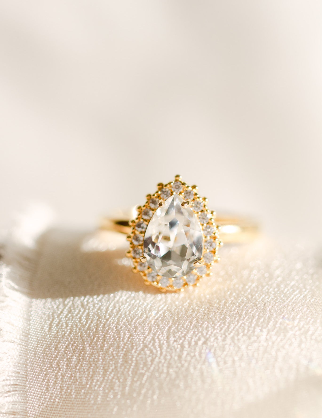 A gold-colored band ring with a central clear stone surrounded by smaller crystals, presented on a white fabric surface.