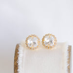 A pair of gold-colored stud earrings with sparkling crystals displayed on a white cushion.