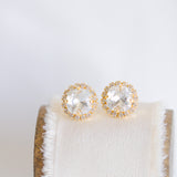 A pair of gold-colored stud earrings with sparkling crystals displayed on a white cushion.