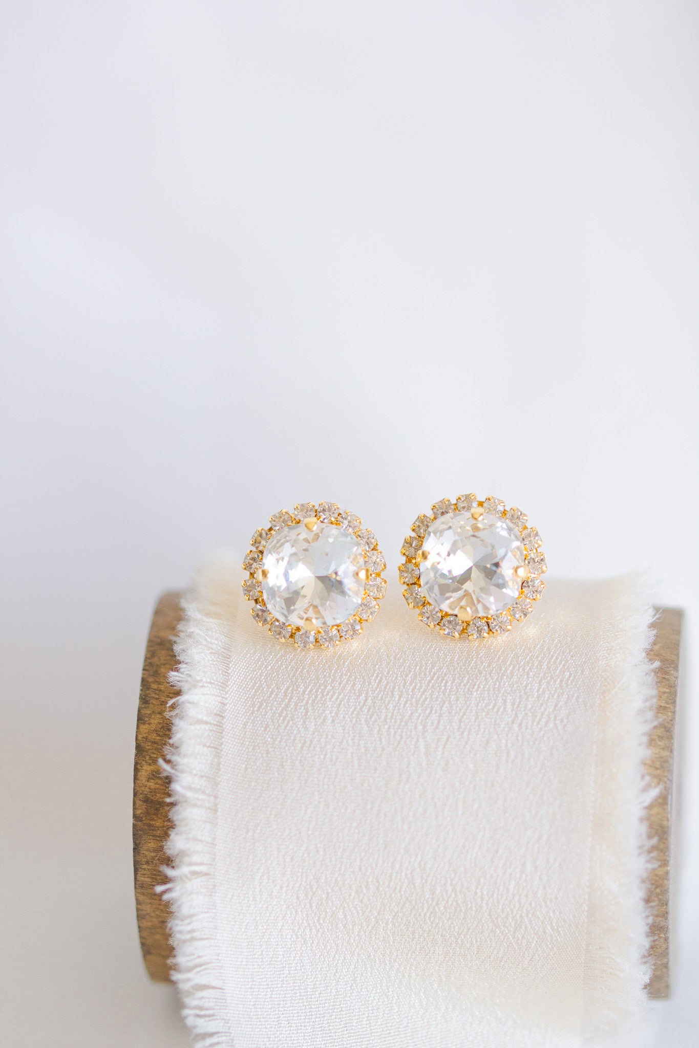A pair of gold-colored stud earrings with sparkling crystals displayed on a white cushion.