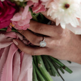 A hand holding a ring with a central oval stone surrounded by a halo of crystals, with a adjustable band. The ring is gold-plated and the central stone is highlighted by the halo of crystals.
