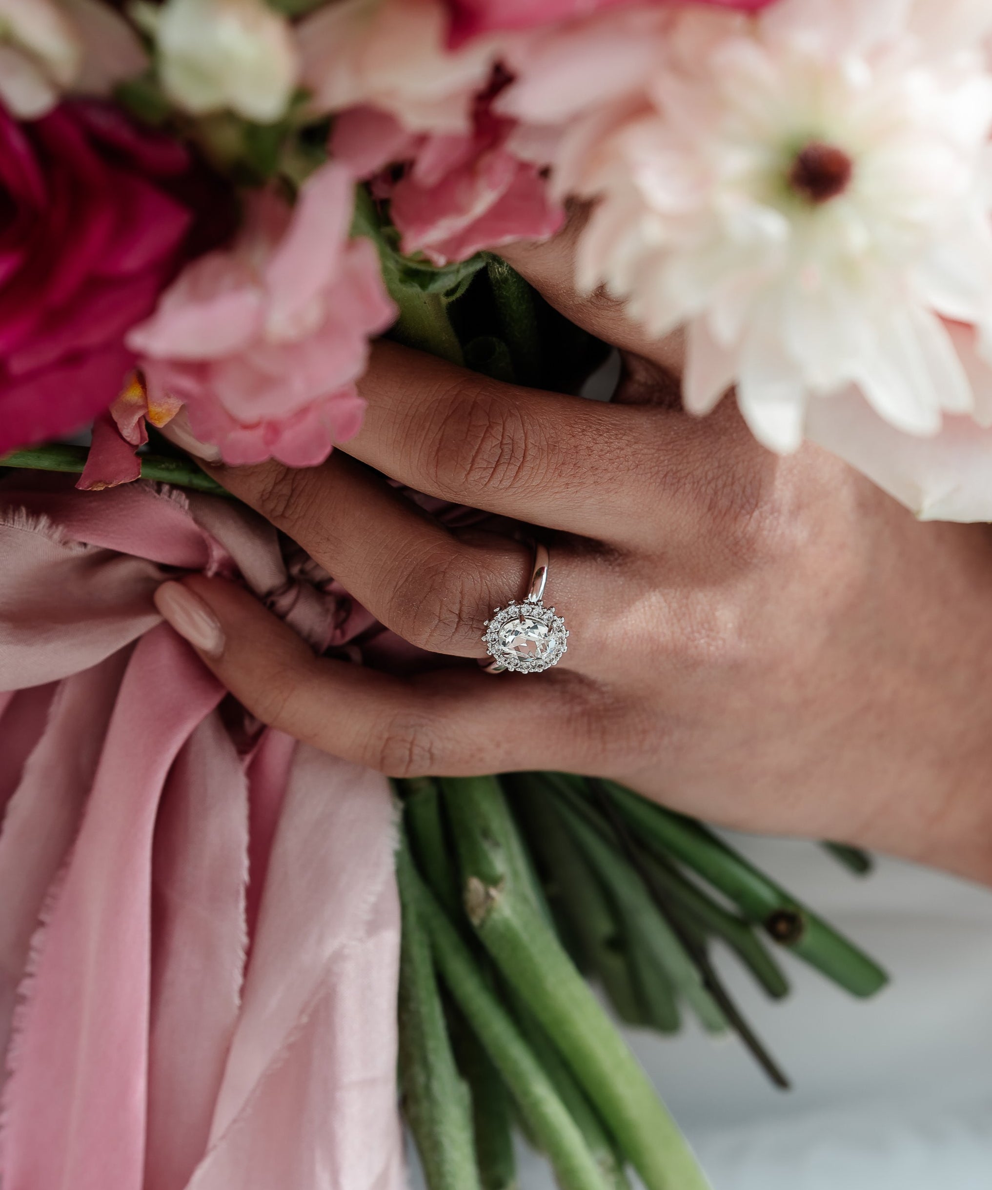 A hand holding a ring with a central oval stone surrounded by a halo of crystals, with a adjustable band. The ring is gold-plated and the central stone is highlighted by the halo of crystals.