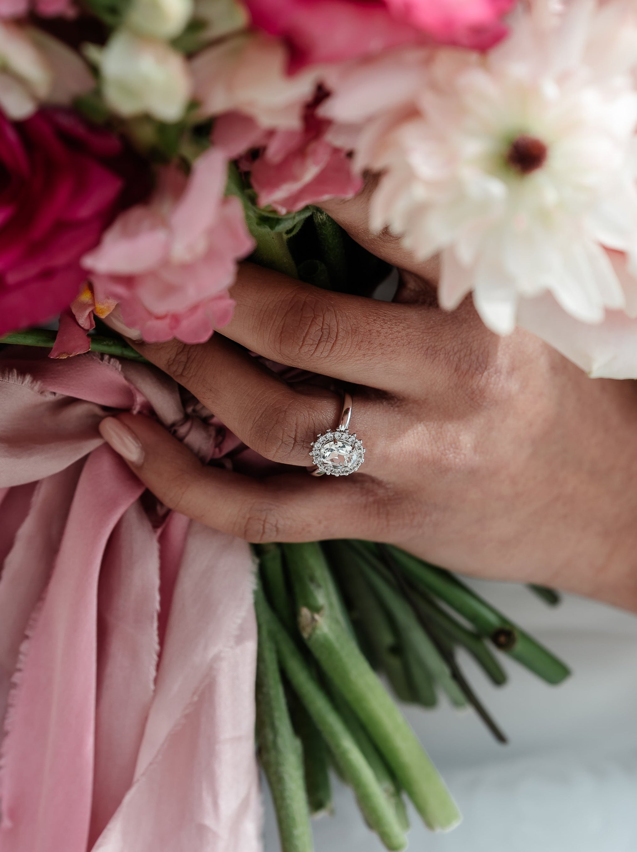 A hand holding a ring with a central oval stone surrounded by a halo of crystals, with a adjustable band. The ring is gold-plated and the central stone is highlighted by the halo of crystals.