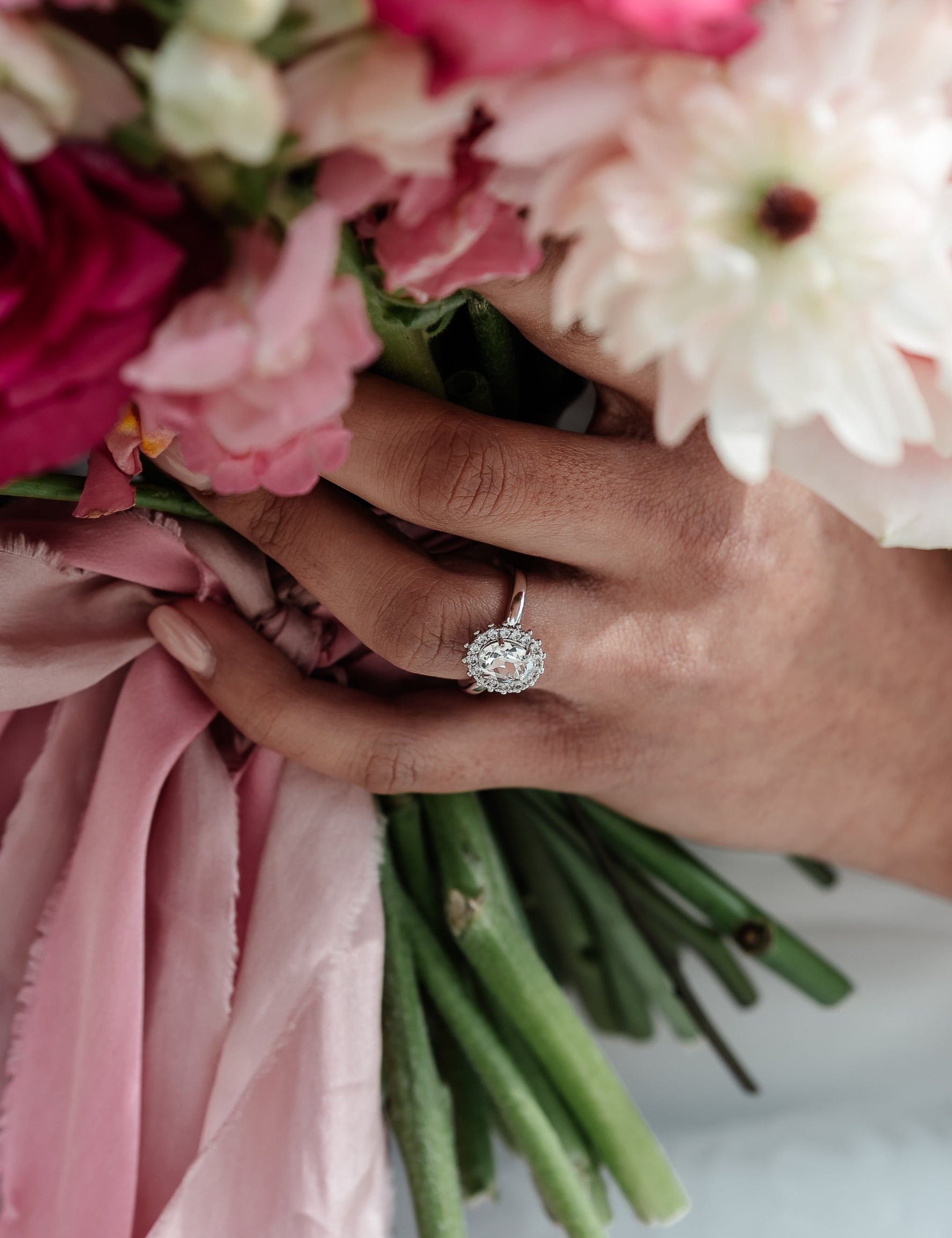 A hand holding a ring with a central oval stone surrounded by a halo of crystals, with a adjustable band. The ring is gold-plated and the central stone is highlighted by the halo of crystals.