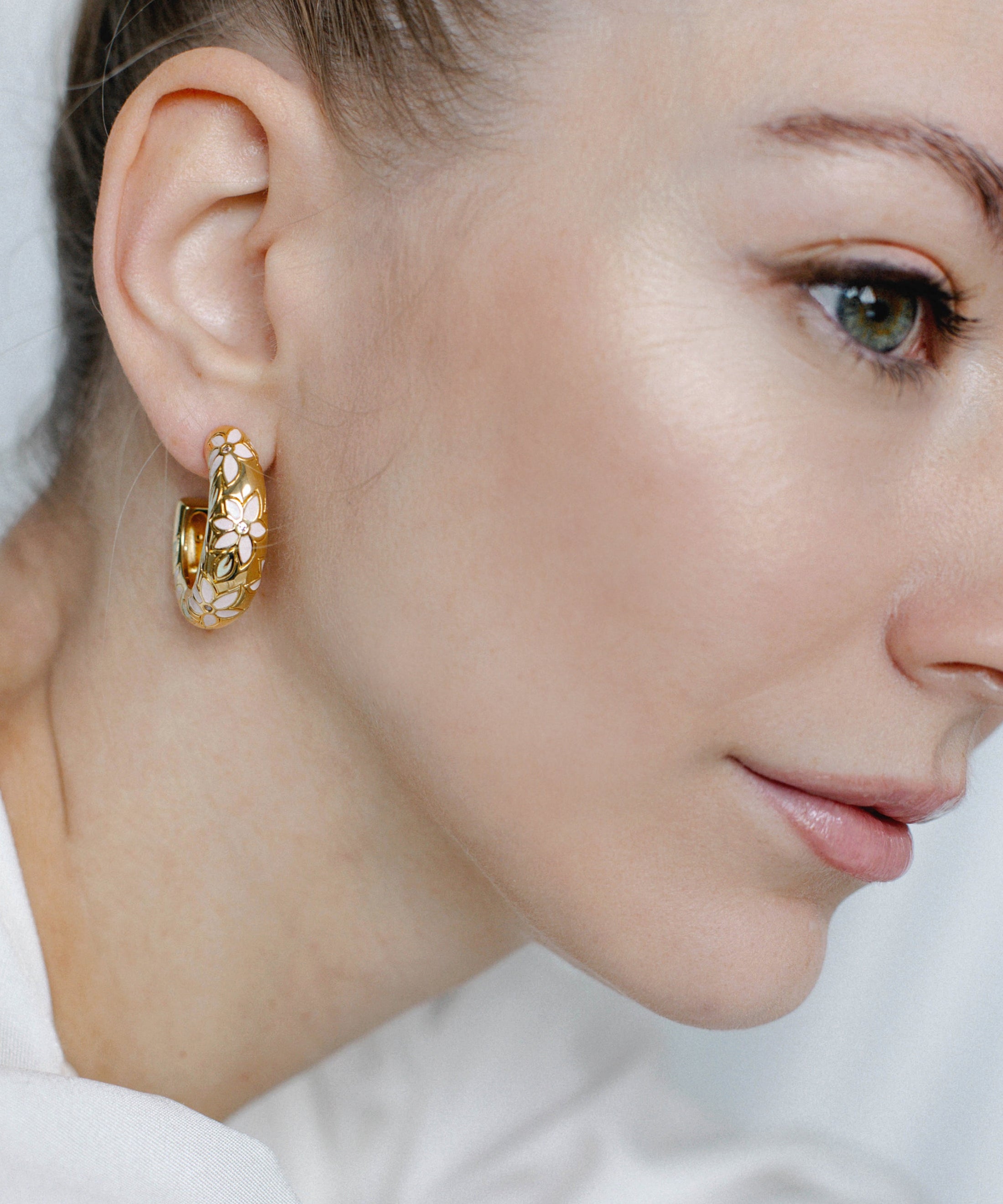 A close-up image of a woman's ear wearing a gold-toned floral hoop earring with hand-painted pastel flowers and crystal accents.