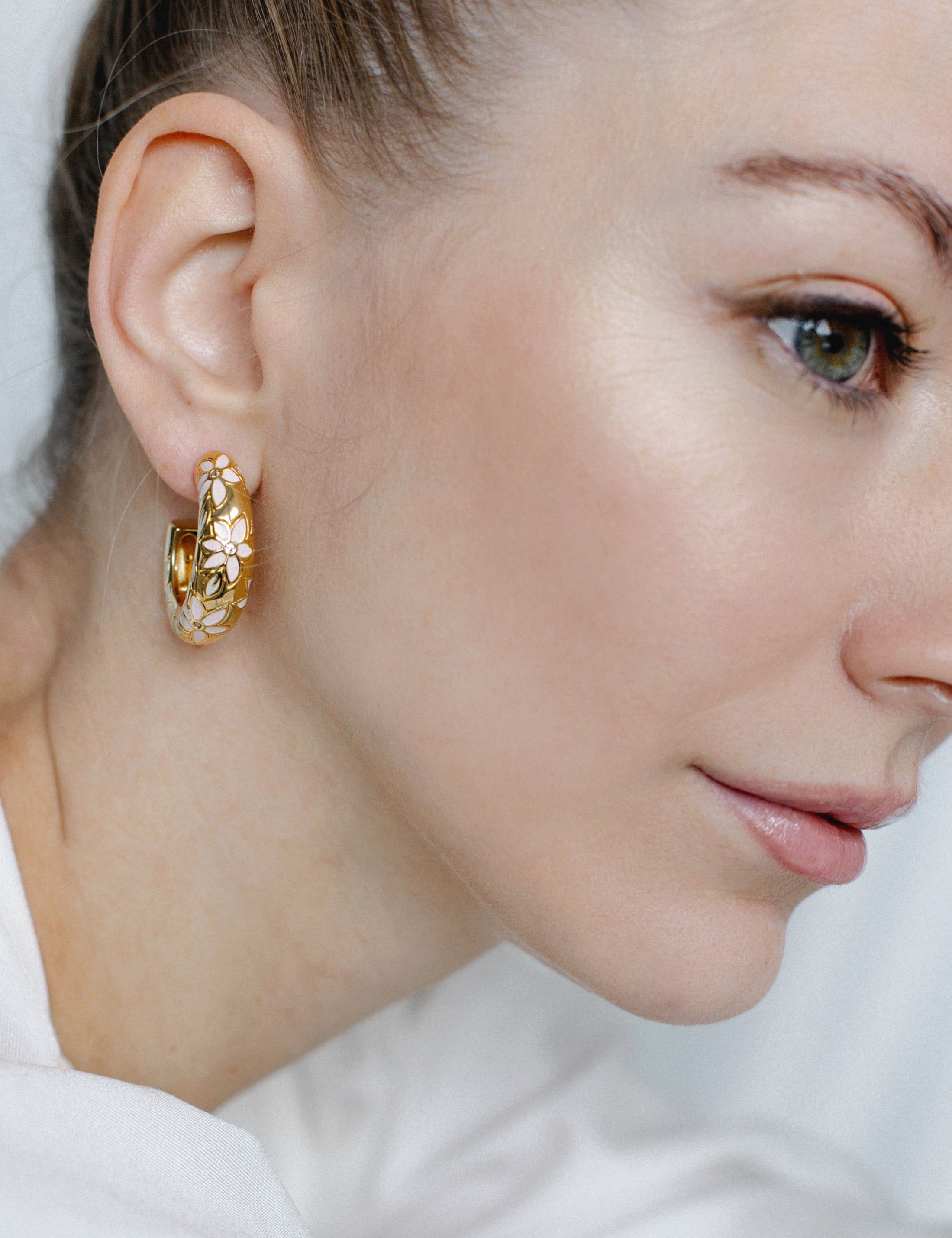 A close-up image of a woman's ear wearing a gold-toned floral hoop earring with hand-painted pastel flowers and crystal accents.