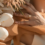 A person's hand wearing a gold ring with a central oval stone surrounded by a halo of crystals, accompanied by flowers.