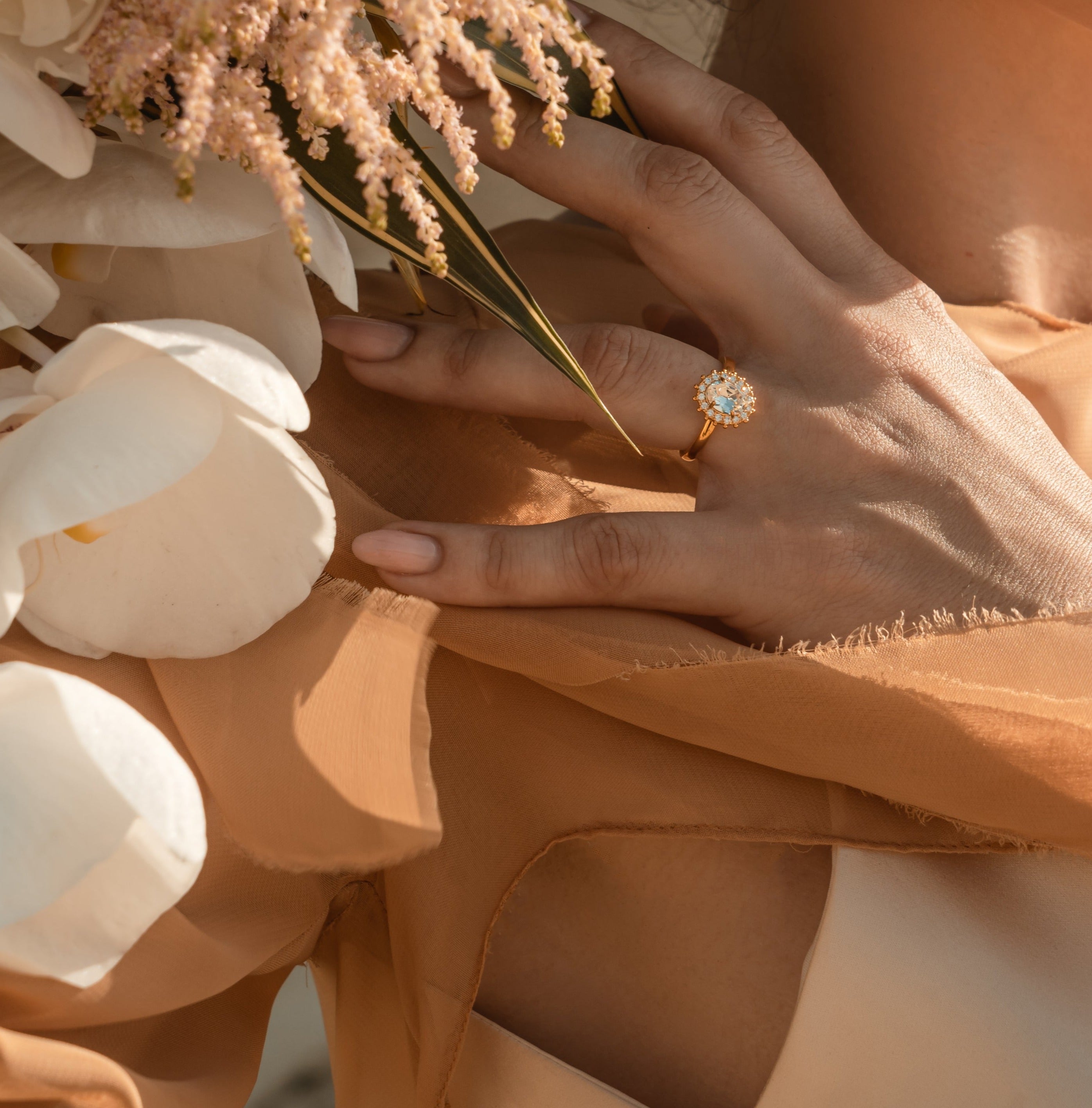 A person's hand wearing a gold ring with a central oval stone surrounded by a halo of crystals, accompanied by flowers.