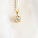 A gold-colored necklace with a round pendant adorned in crystals, displayed against a white fabric background.