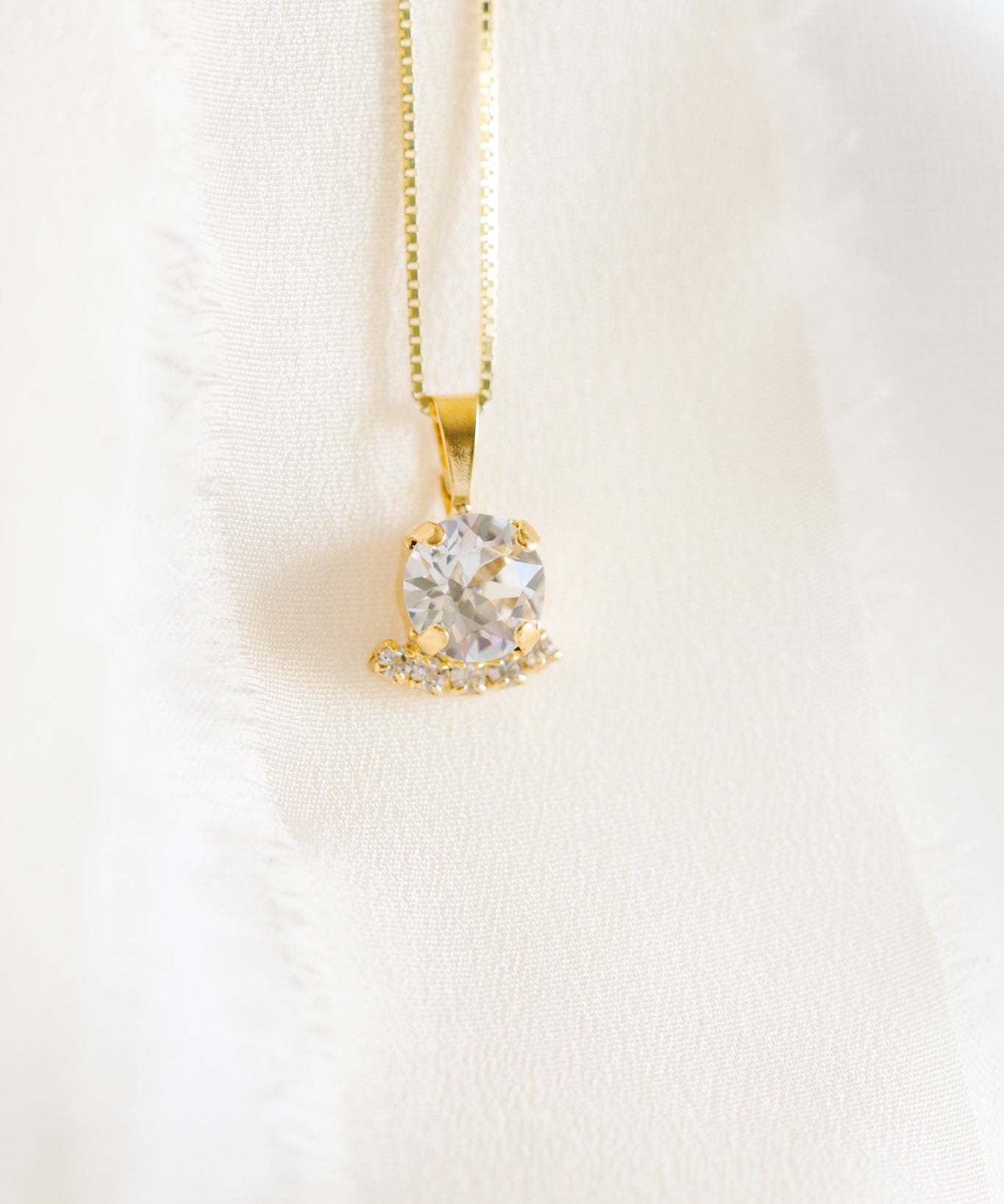 A gold-colored necklace with a round pendant adorned in crystals, displayed against a white fabric background.