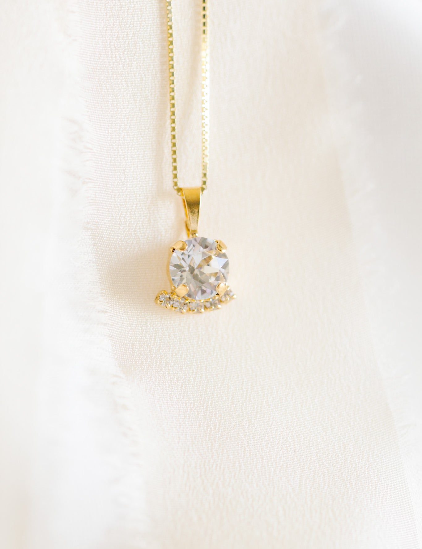 A gold-colored necklace with a round pendant adorned in crystals, displayed against a white fabric background.