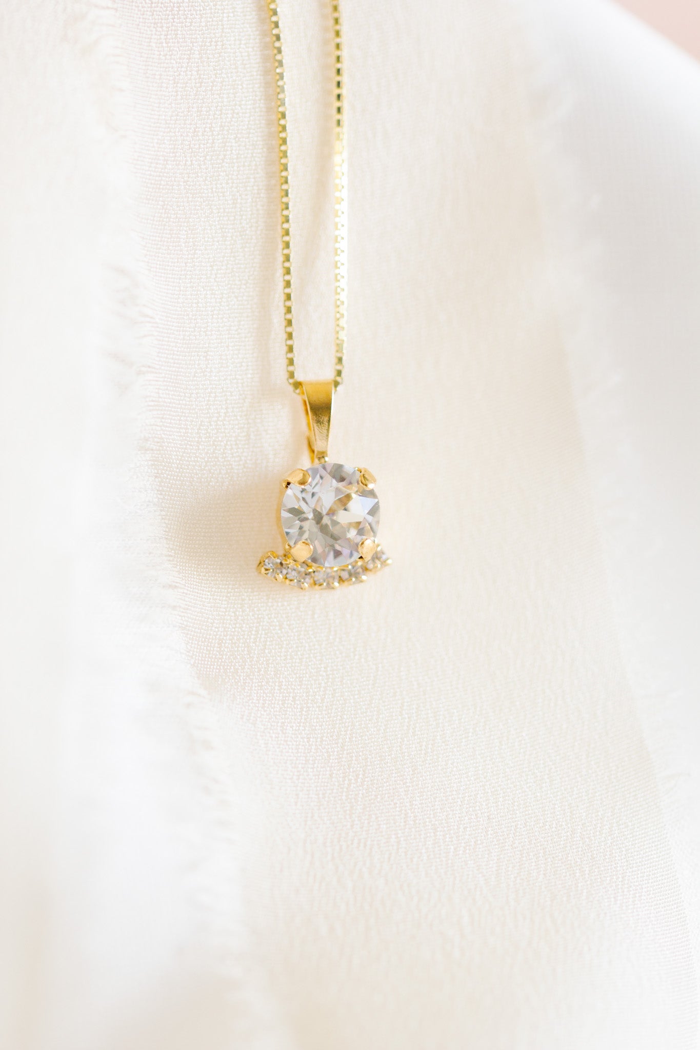 A gold-colored necklace with a round pendant adorned in crystals, displayed against a white fabric background.