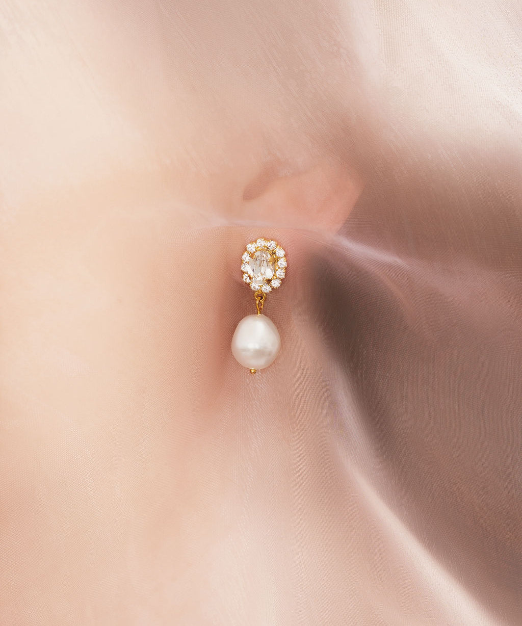 A close-up image of a woman's ear wearing a gold-plated earring with a baroque pearl and a sparkling crystal atop.