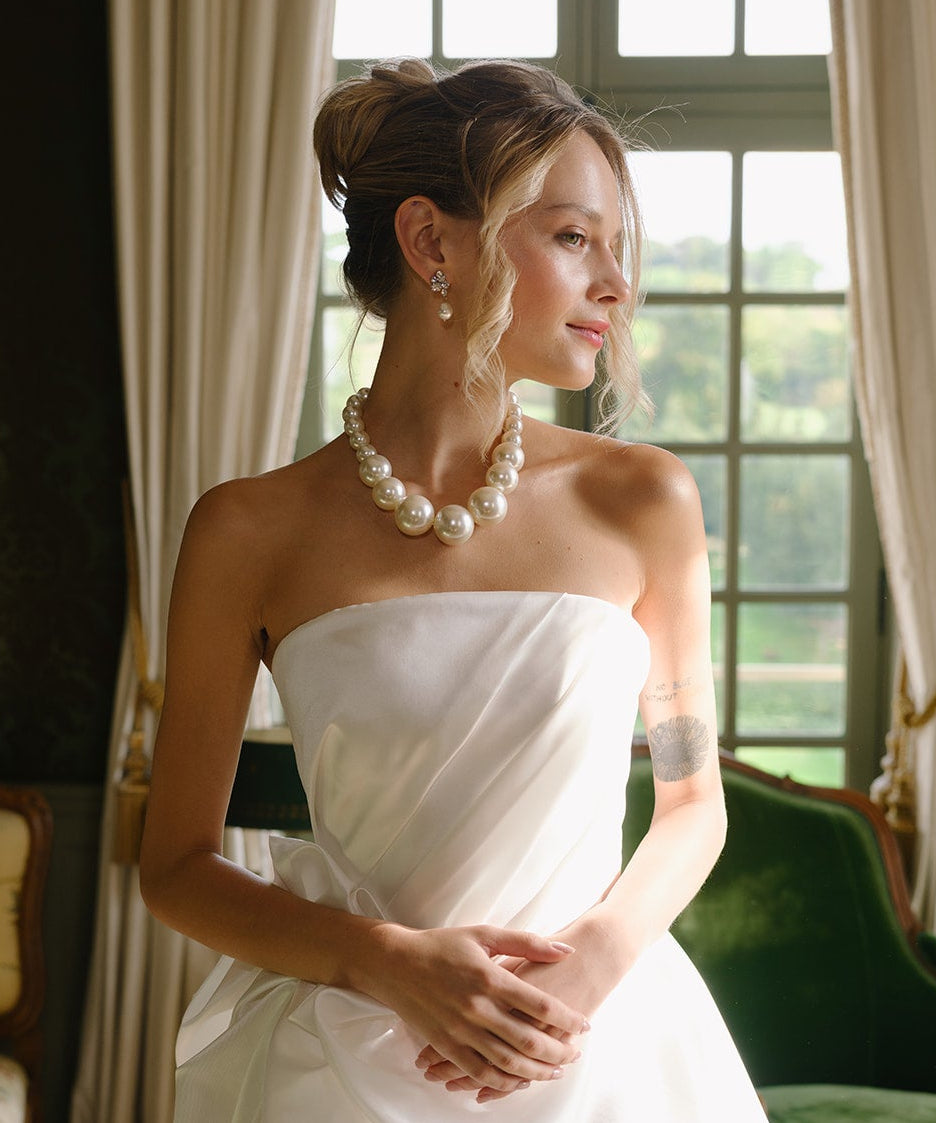 Woman in a white strapless dress with a pearl necklace and wedding earrings standing in a room with green walls and a large window.