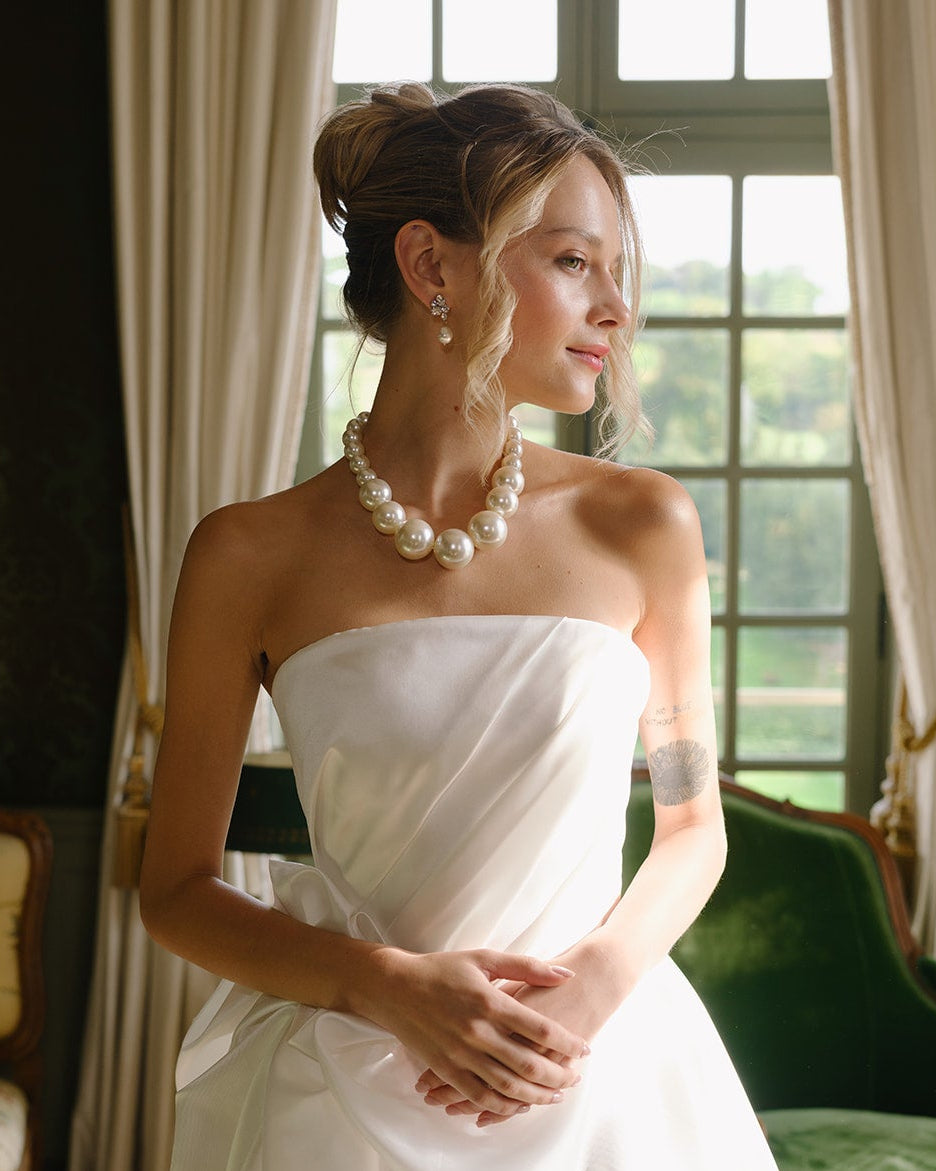 Woman in a white strapless dress with a pearl necklace and wedding earrings standing in a room with green walls and a large window.