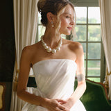 Woman in a white strapless dress with a pearl necklace and wedding earrings standing in a room with green walls and a large window.