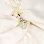 A gold-plated necklace with a cushion-cut stone centerpiece surrounded by crystals, displayed on a white fabric surface.