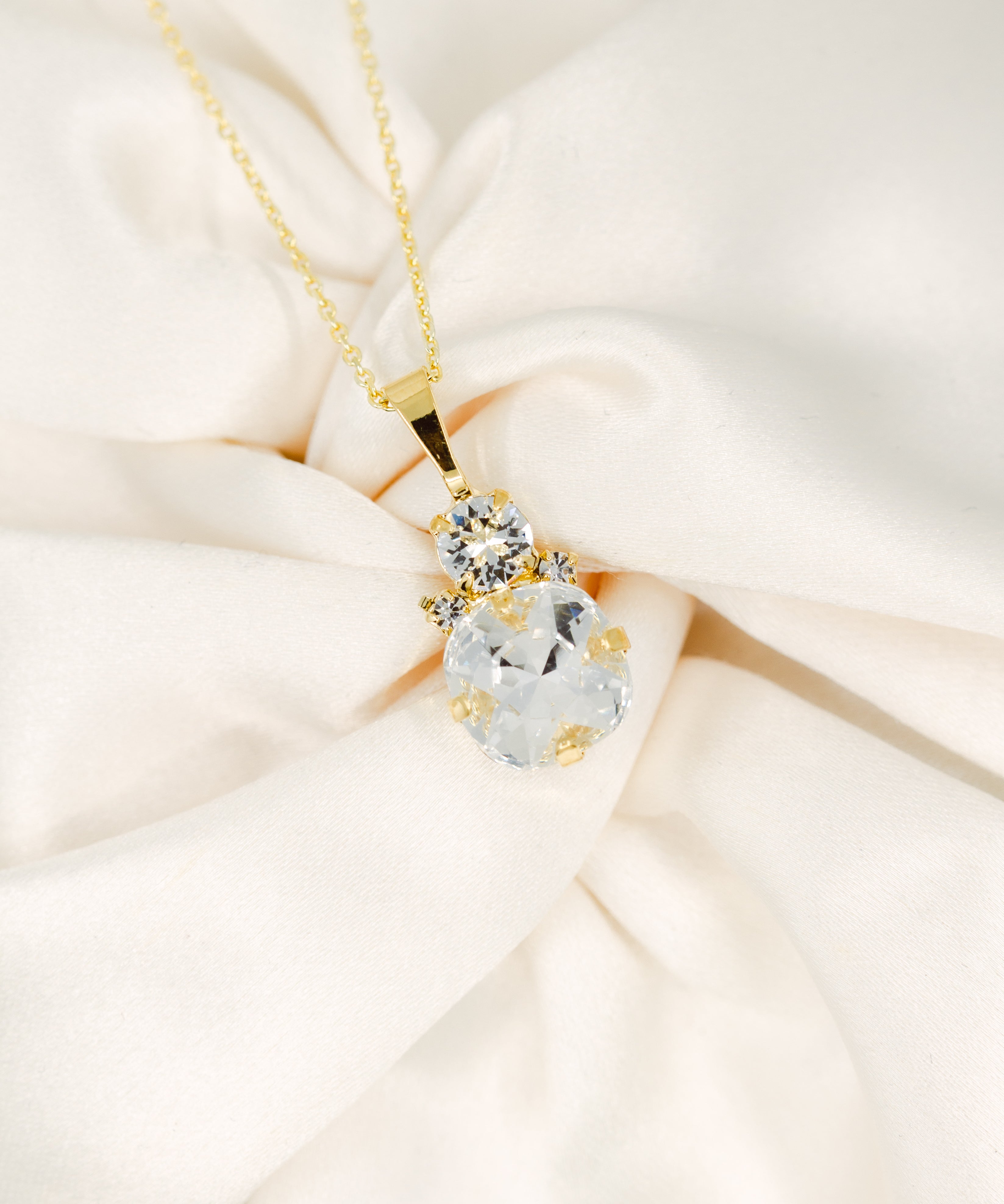 A gold-plated necklace with a cushion-cut stone centerpiece surrounded by crystals, displayed on a white fabric surface.