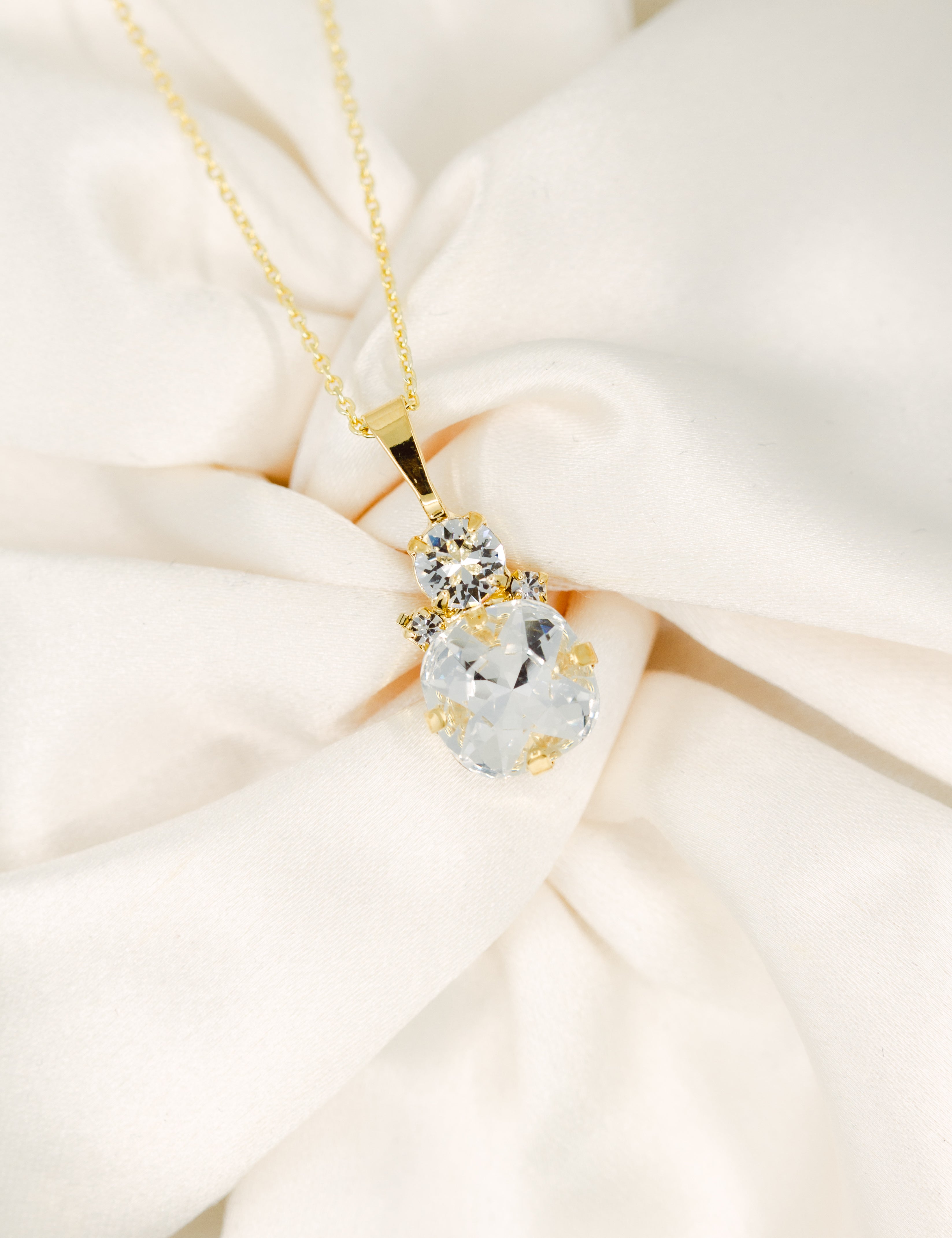 A gold-plated necklace with a cushion-cut stone centerpiece surrounded by crystals, displayed on a white fabric surface.