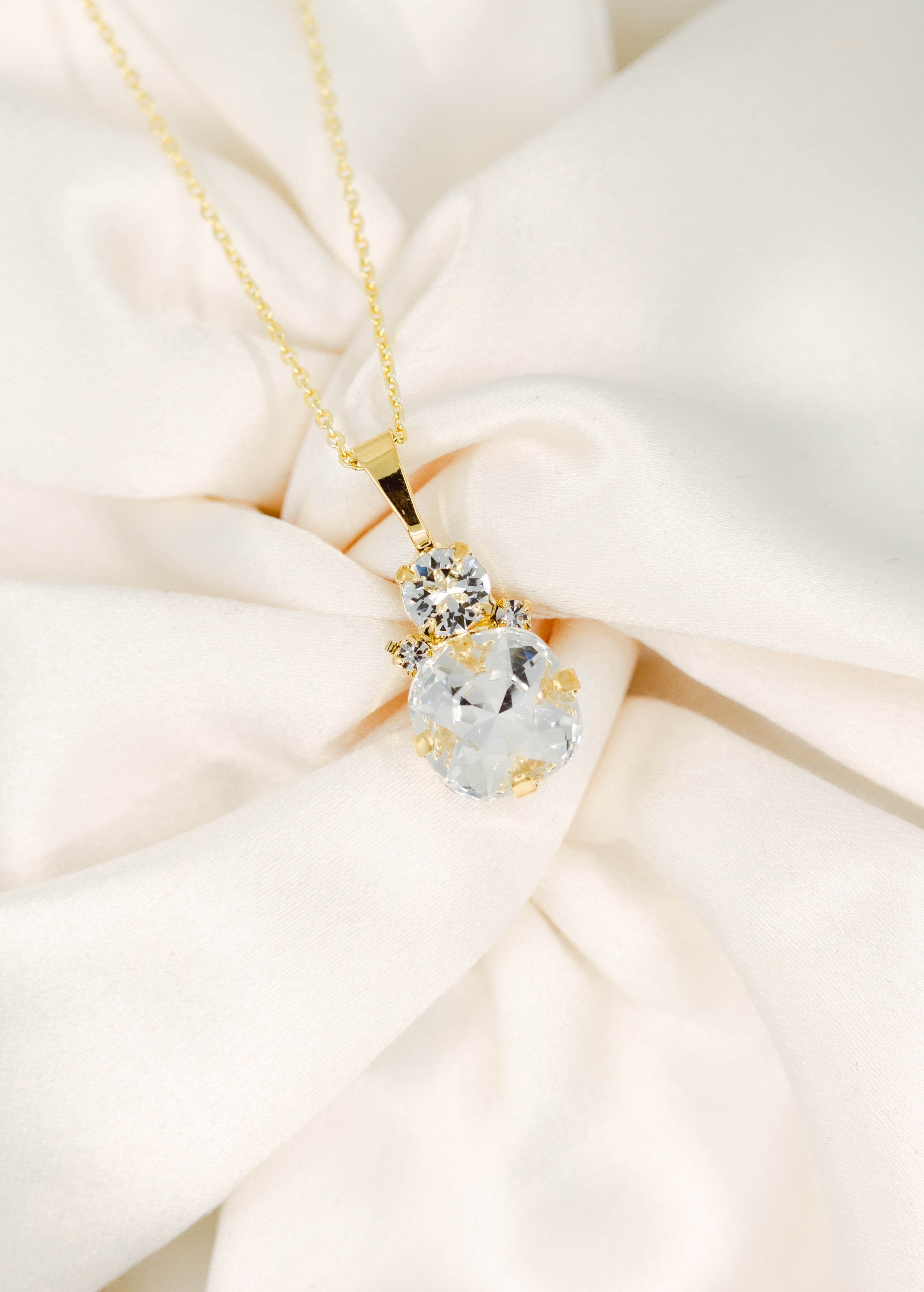 A gold-plated necklace with a cushion-cut stone centerpiece surrounded by crystals, displayed on a white fabric surface.