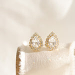 A pair of gold-plated earrings featuring a teardrop shape with a halo of smaller crystals around the main crystal, displayed on a white cushion.