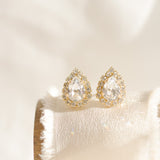 A pair of gold-plated earrings featuring a teardrop shape with a halo of smaller crystals around the main crystal, displayed on a white cushion.