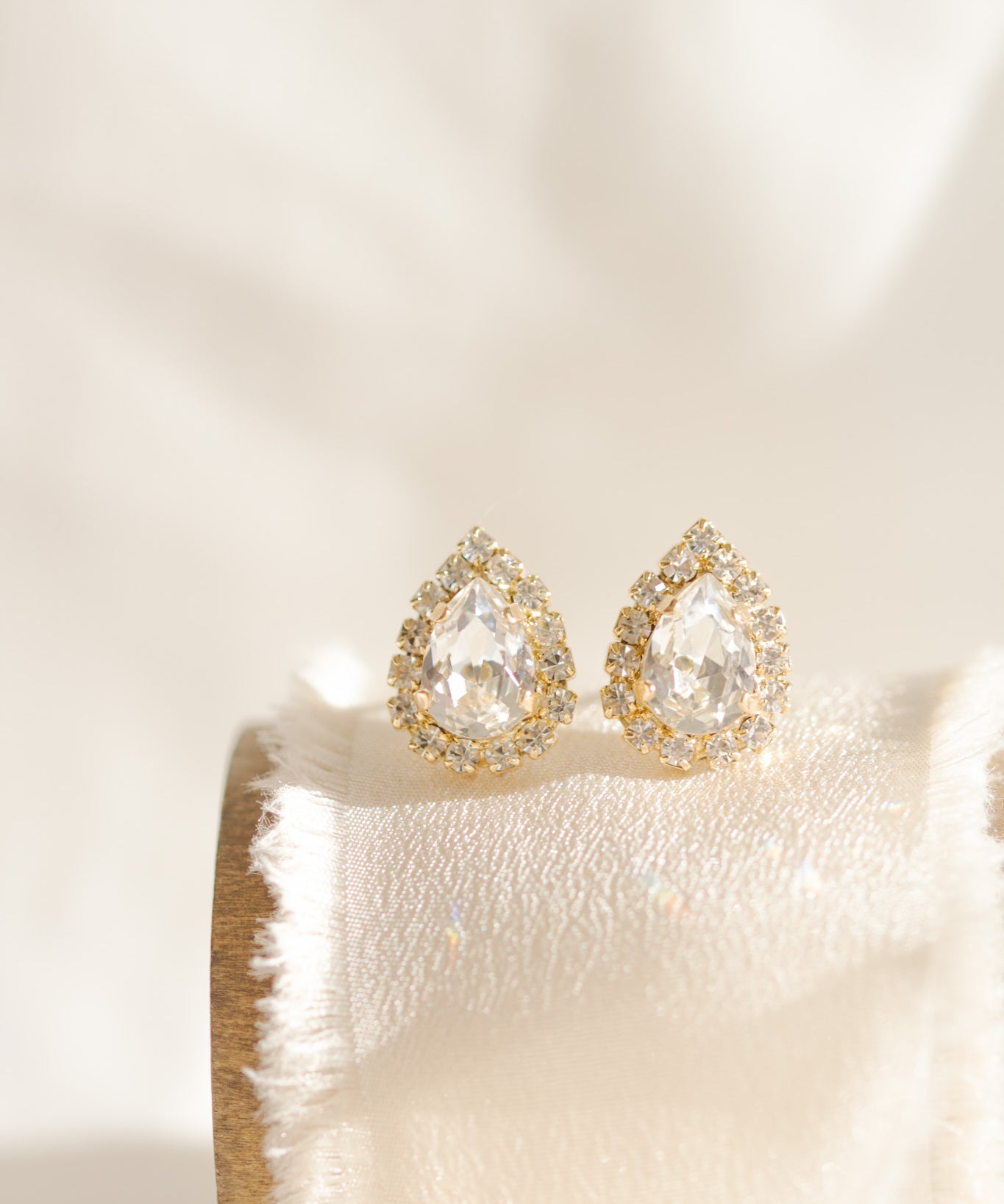 A pair of gold-plated earrings featuring a teardrop shape with a halo of smaller crystals around the main crystal, displayed on a white cushion.