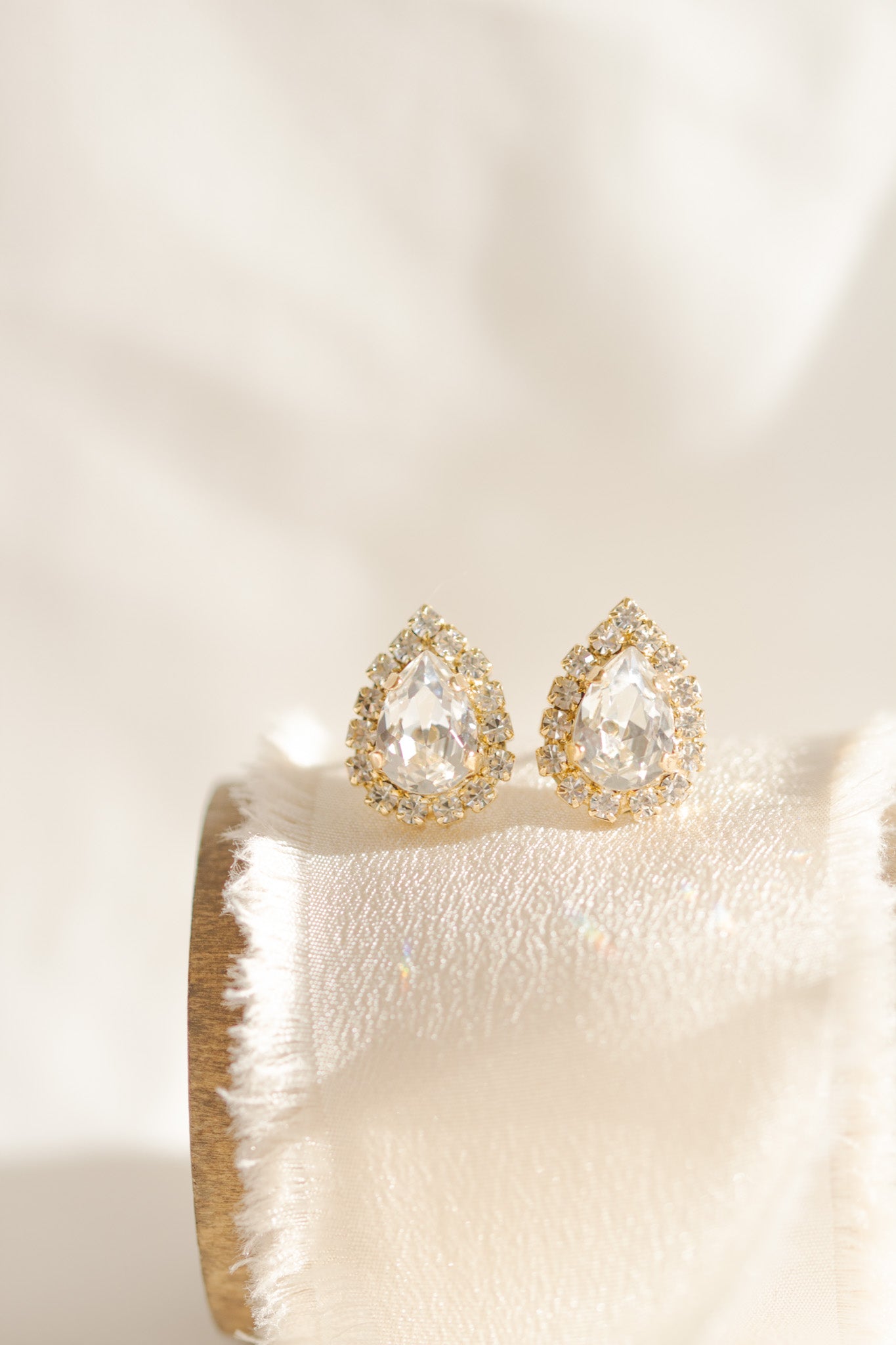 A pair of gold-plated earrings featuring a teardrop shape with a halo of smaller crystals around the main crystal, displayed on a white cushion.