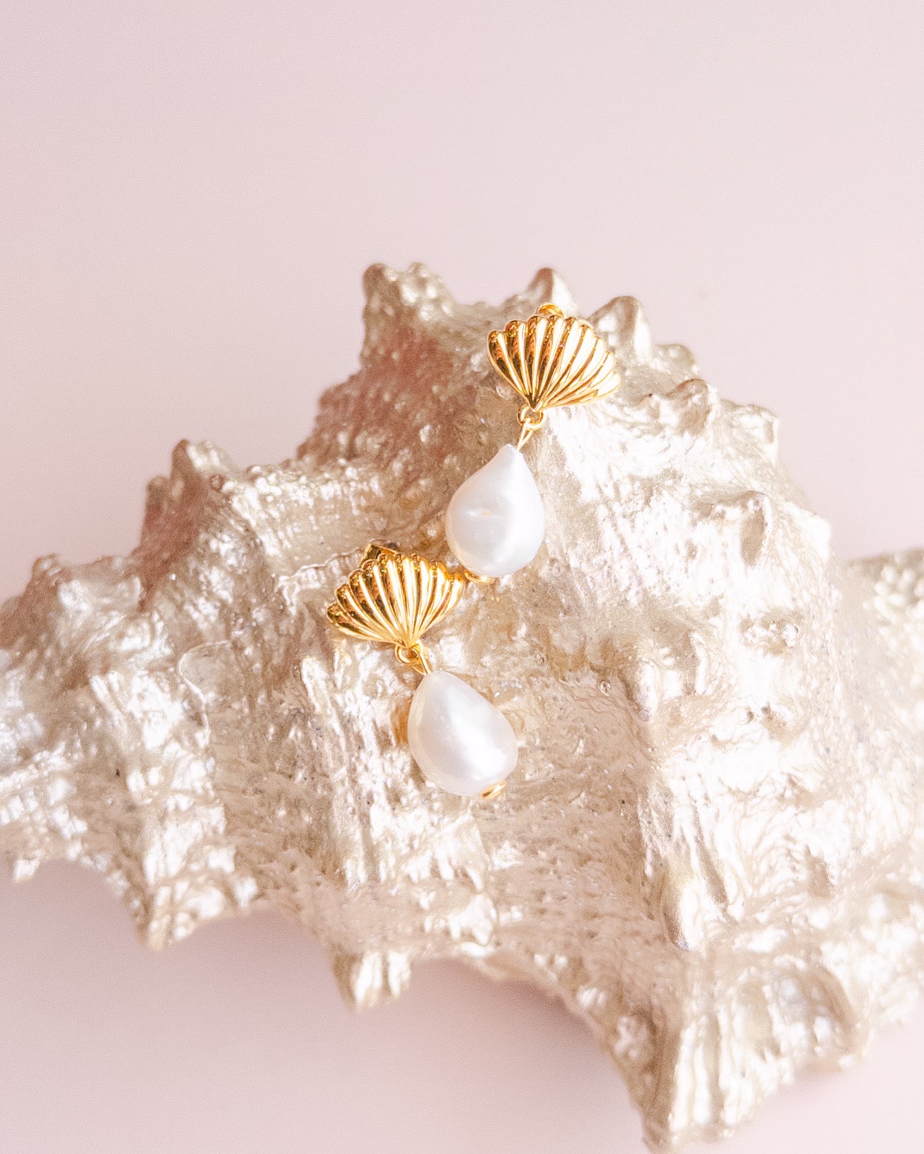 A pair of gold-plated scallop earrings with freshwater pearls displayed on a seashell by Sarah Gauci