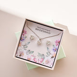 A jewelry set including a necklace and stud earrings, displayed in a box with a floral pattern and the text 'Thank you for being my Bridesmaid'. The jewelry is plated in gold, and has gold and white crystals.