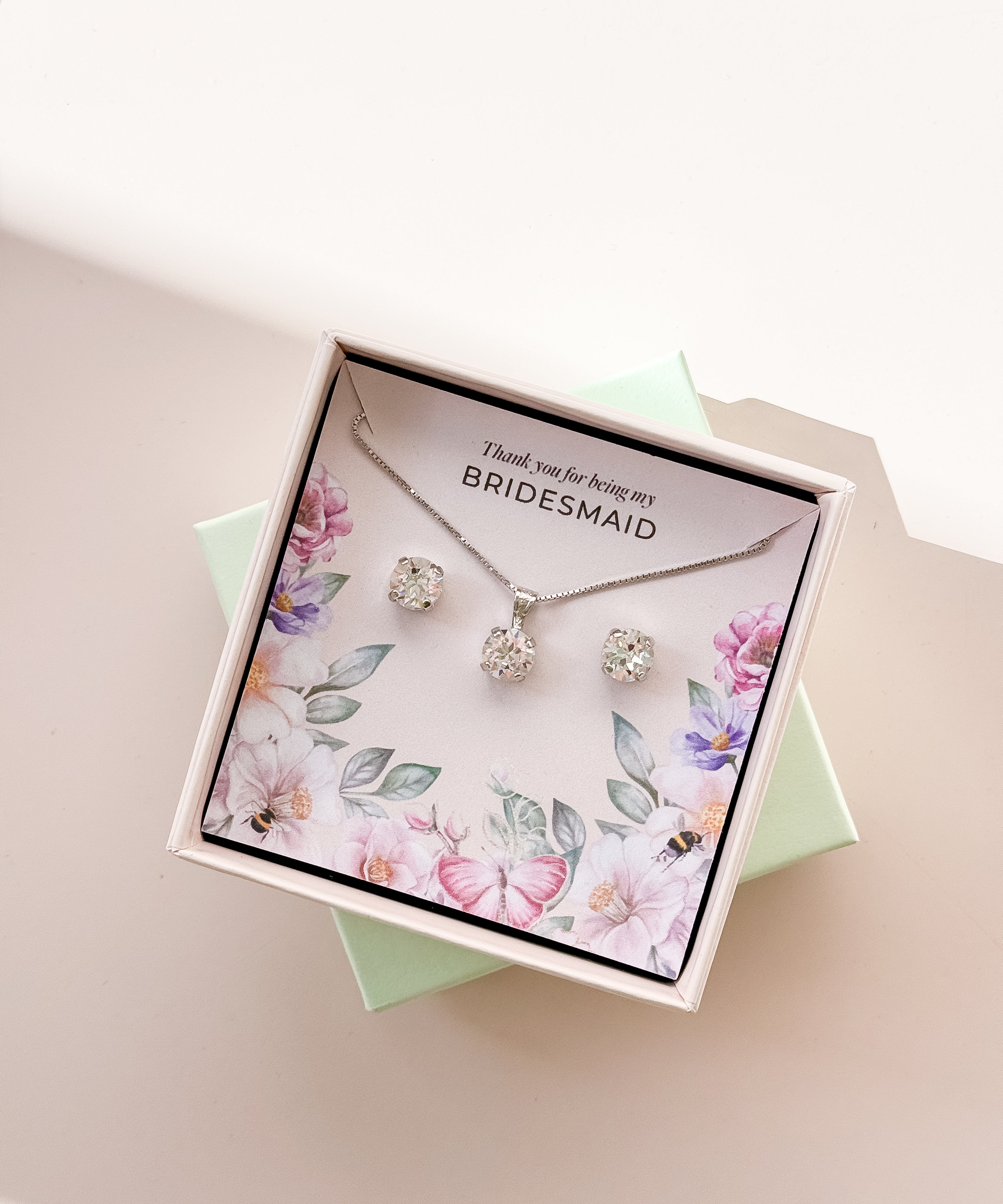 A jewelry set including a necklace and stud earrings, displayed in a box with a floral pattern and the text 'Thank you for being my Bridesmaid'. The jewelry is plated in gold, and has gold and white crystals.
