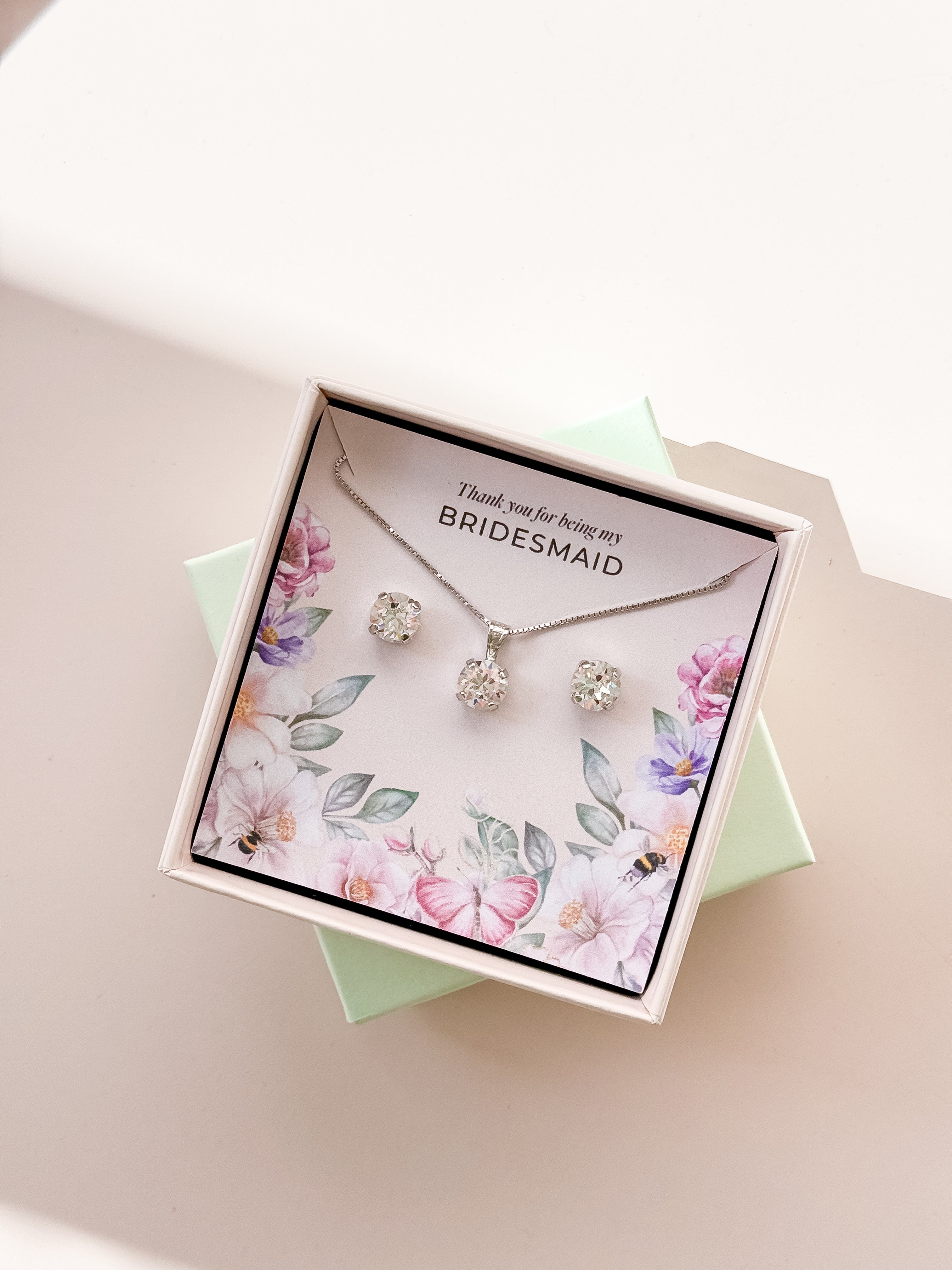 A jewelry set including a necklace and stud earrings, displayed in a box with a floral pattern and the text 'Thank you for being my Bridesmaid'. The jewelry is plated in gold, and has gold and white crystals.
