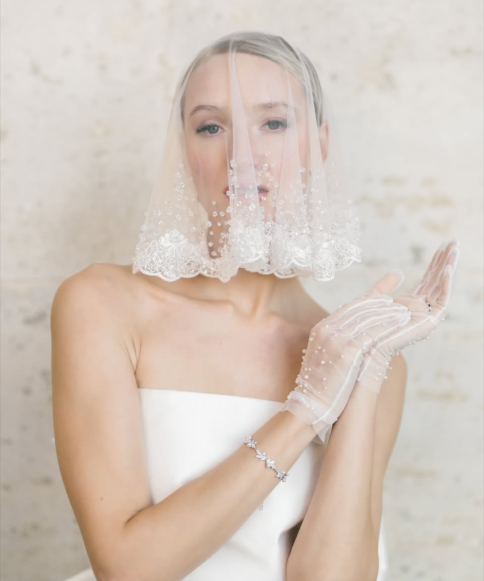 A Bride wearing a white wedding dress, veil, and gloves against a neutral background