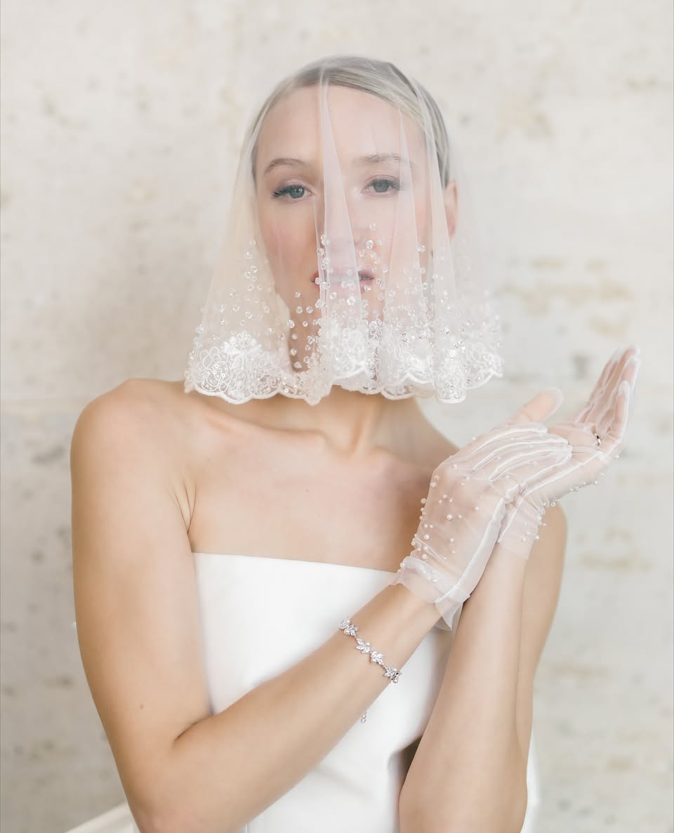A Bride wearing a white wedding dress, veil, and gloves against a neutral background