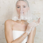 A Bride wearing a white wedding dress, veil, and gloves against a neutral background