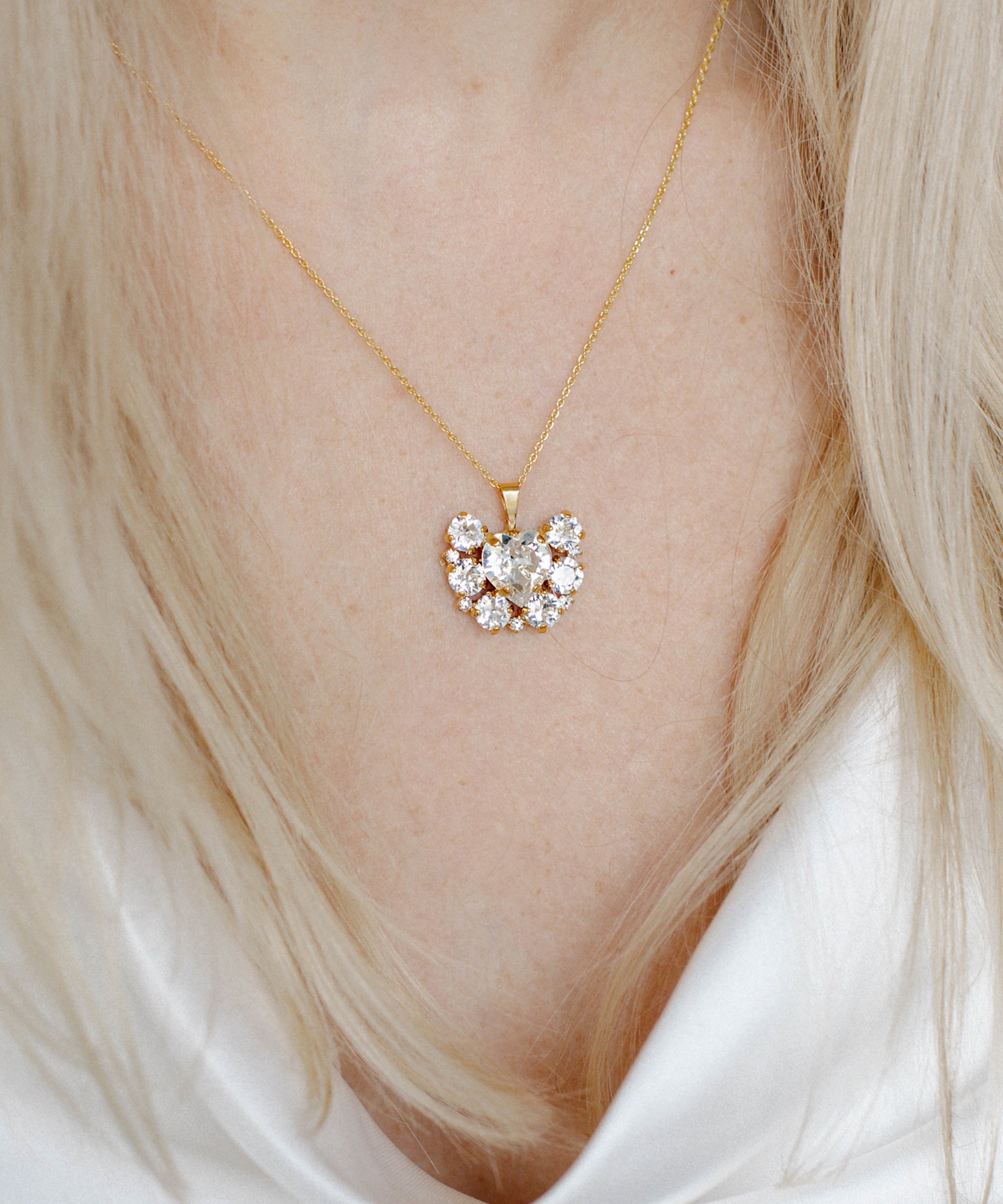 A person wearing a white v-neck top showcasing a gold-plated necklace with a floral pattern and a central heart-shaped crystal pendant.